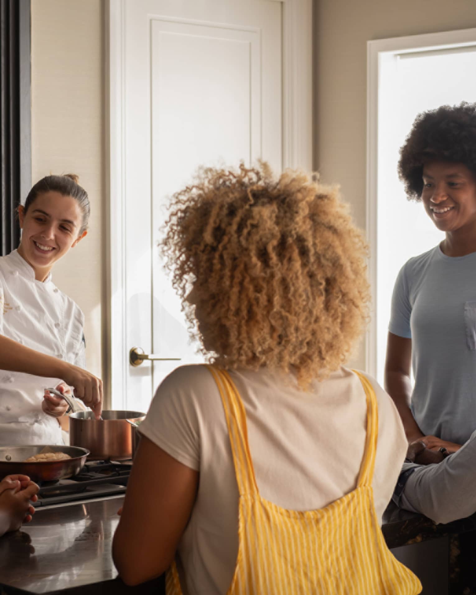 A family of four gathers around a kitchen-island cooktop watching a smiling chef prepare food in copper cookware. 