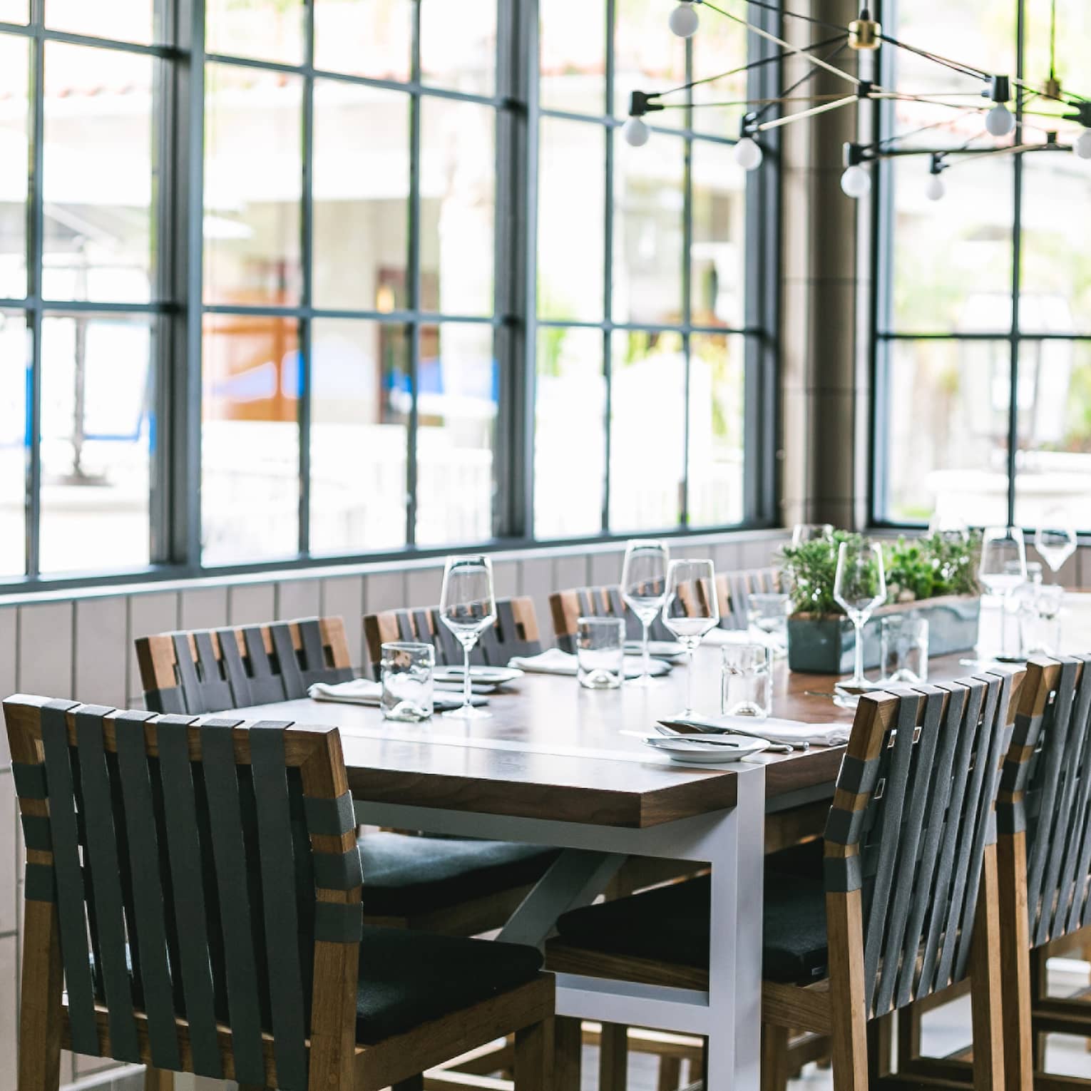 A rectangular wooden dining table with 12 black chairs under an industrial chandelier in a bright room with tiled walls. 