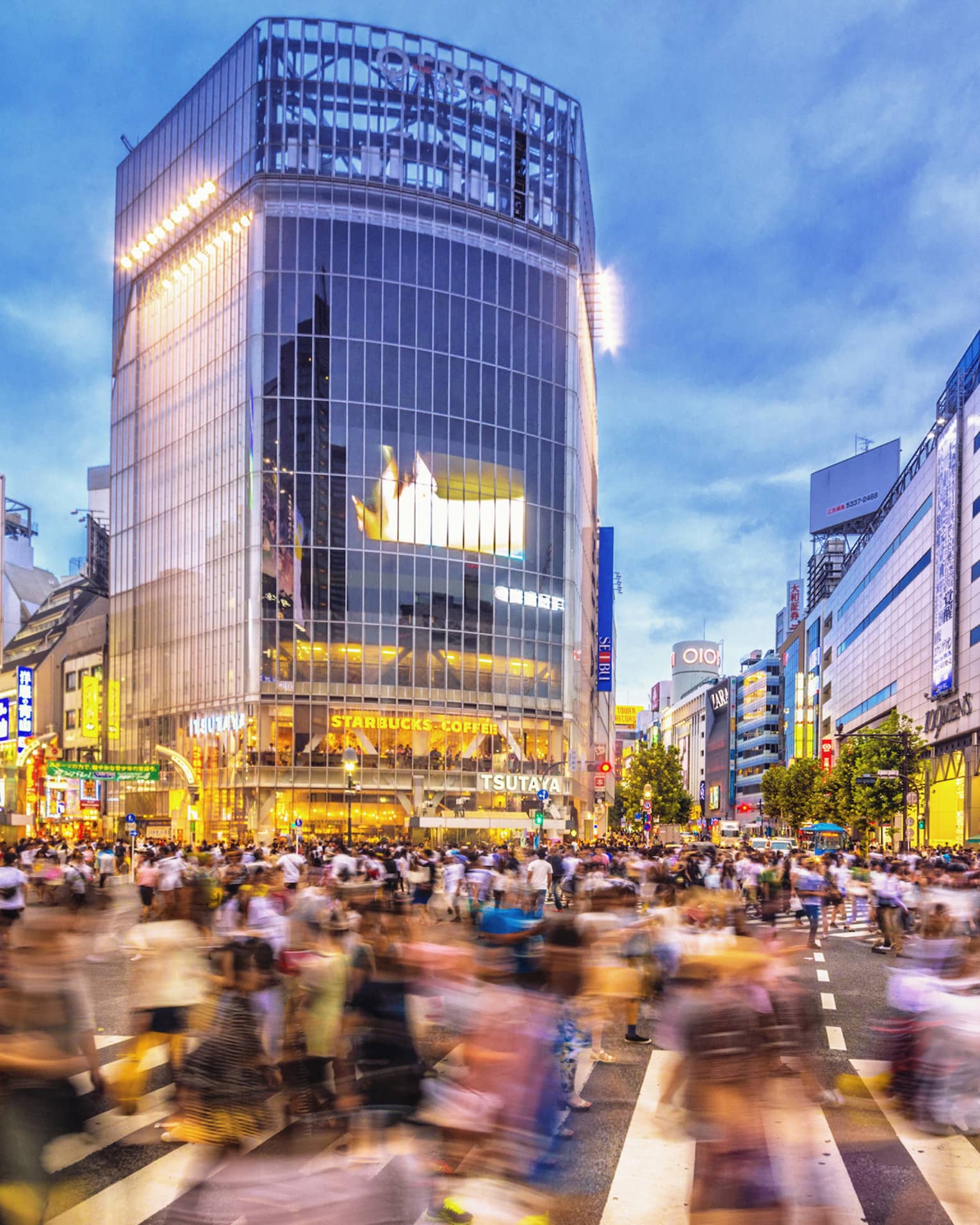 Image of blurry crowds along sidewalk under glass buildings, colourful billboards at night