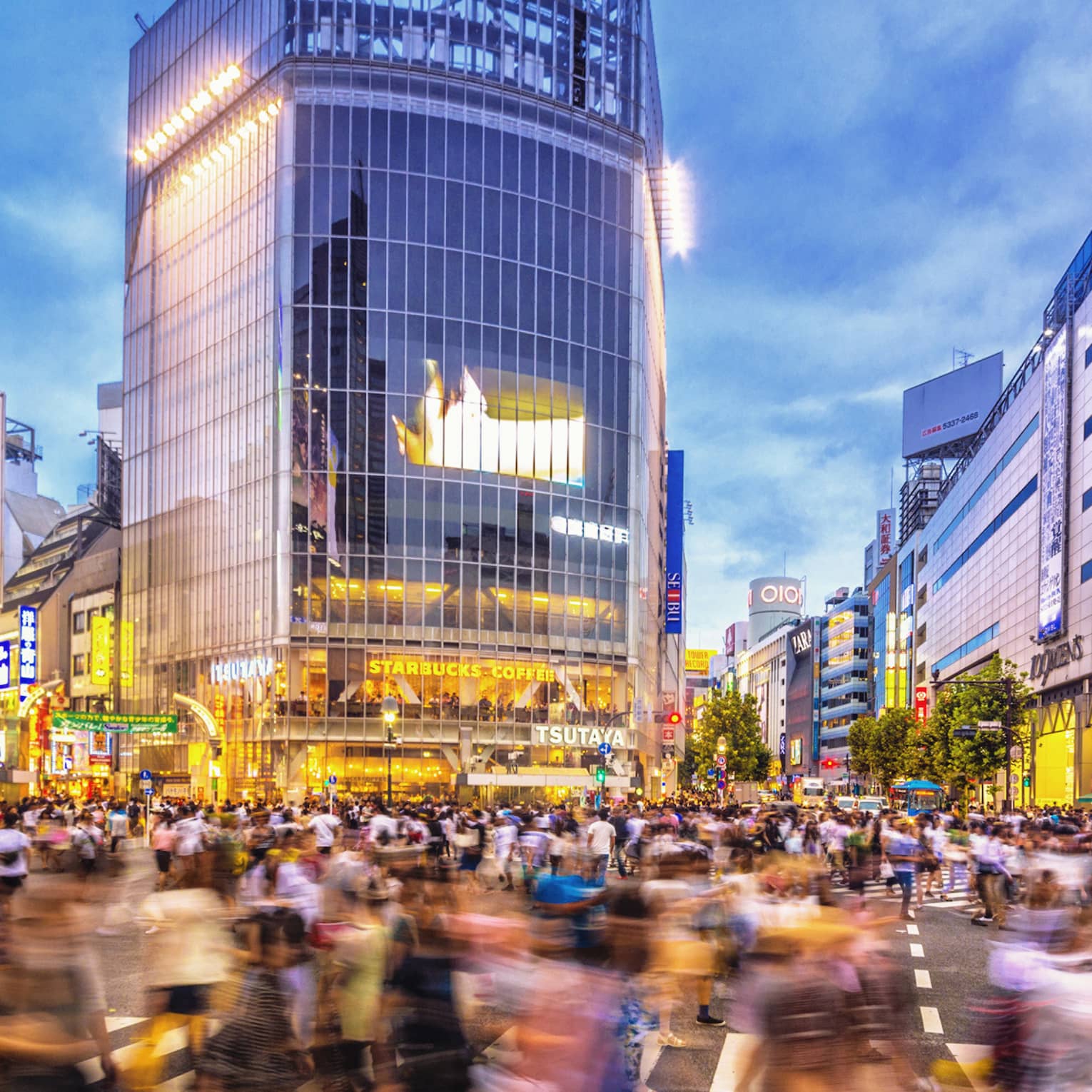 Image of blurry crowds along sidewalk under glass buildings, colourful billboards at night