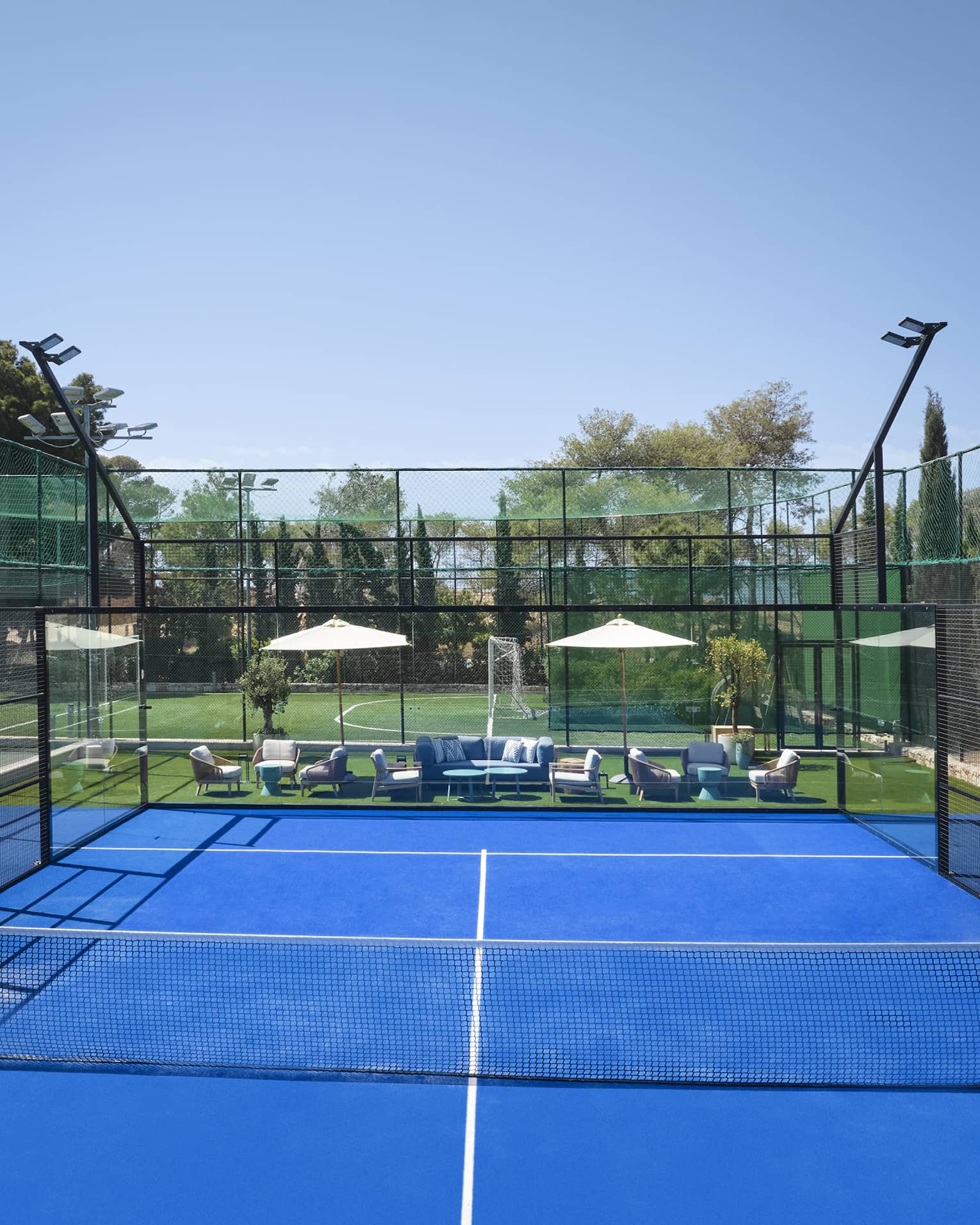 Bright blue padel court surrounded by fencing, white umbrellas and lounge chairs