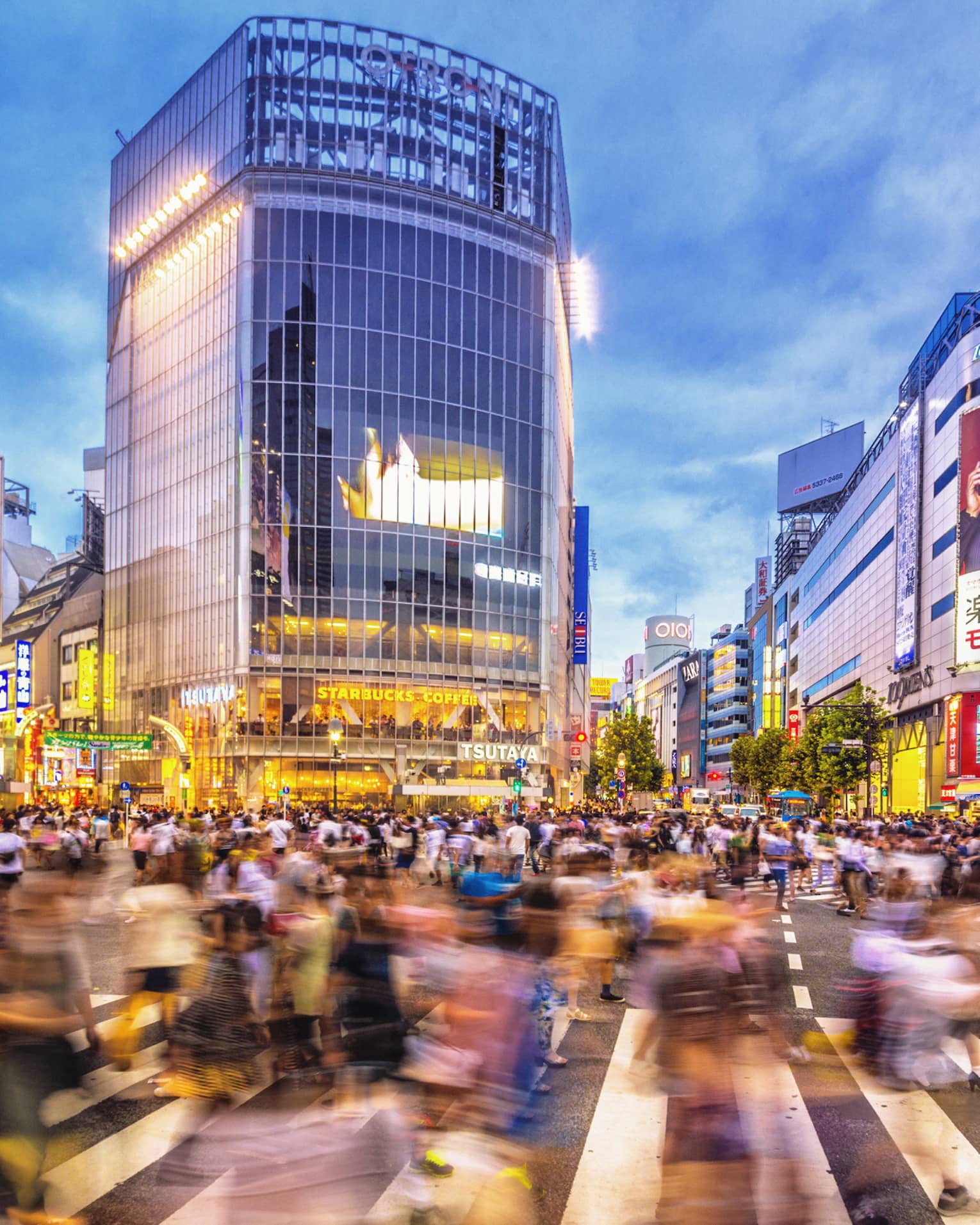 Image of blurry crowds along sidewalk under glass buildings, colourful billboards at night