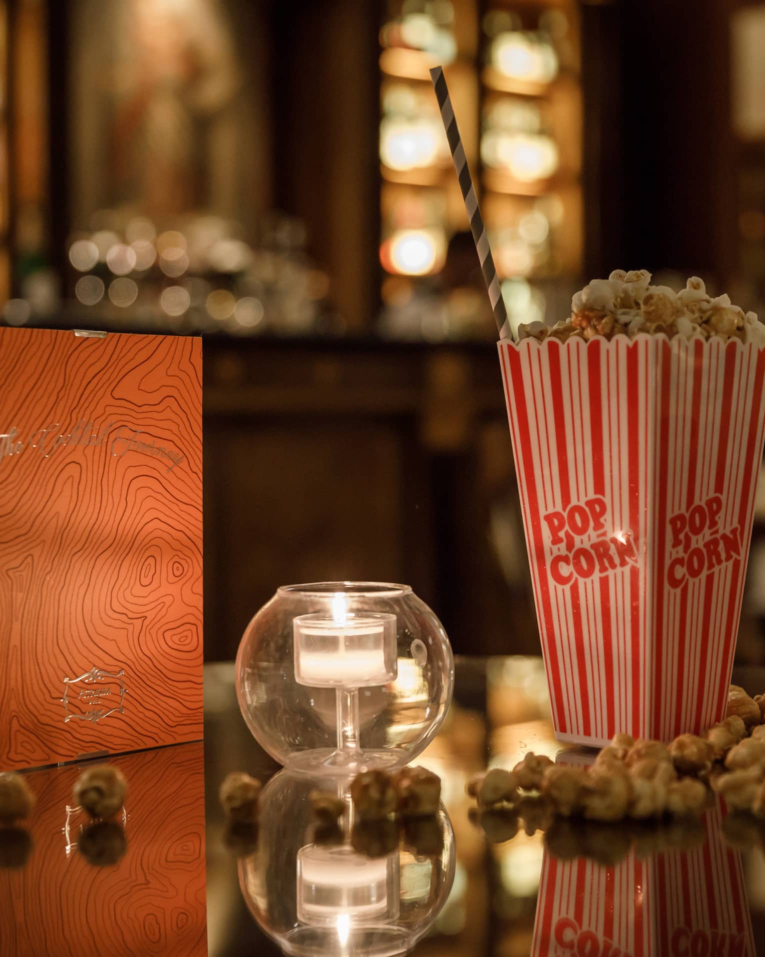 A red and gold box of chocolates, a candle in clear glass and a tall box of popcorn on a shiny table reflecting warm lights.