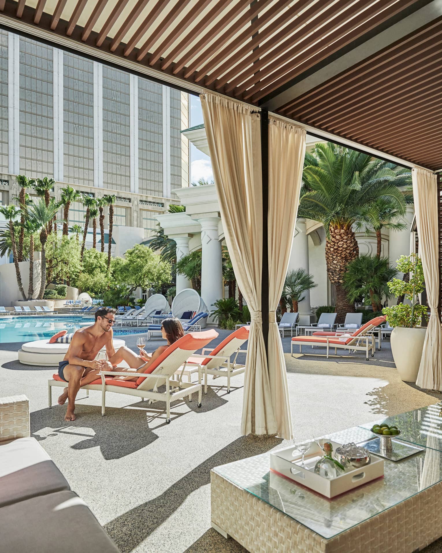 Couple on patio chairs between outdoor swimming pool and cabana seating area 