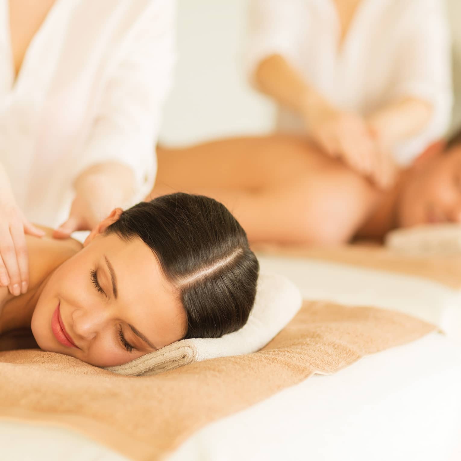 Woman and man lie side-by-side on massage tables as spa staff massage their bare shoulders