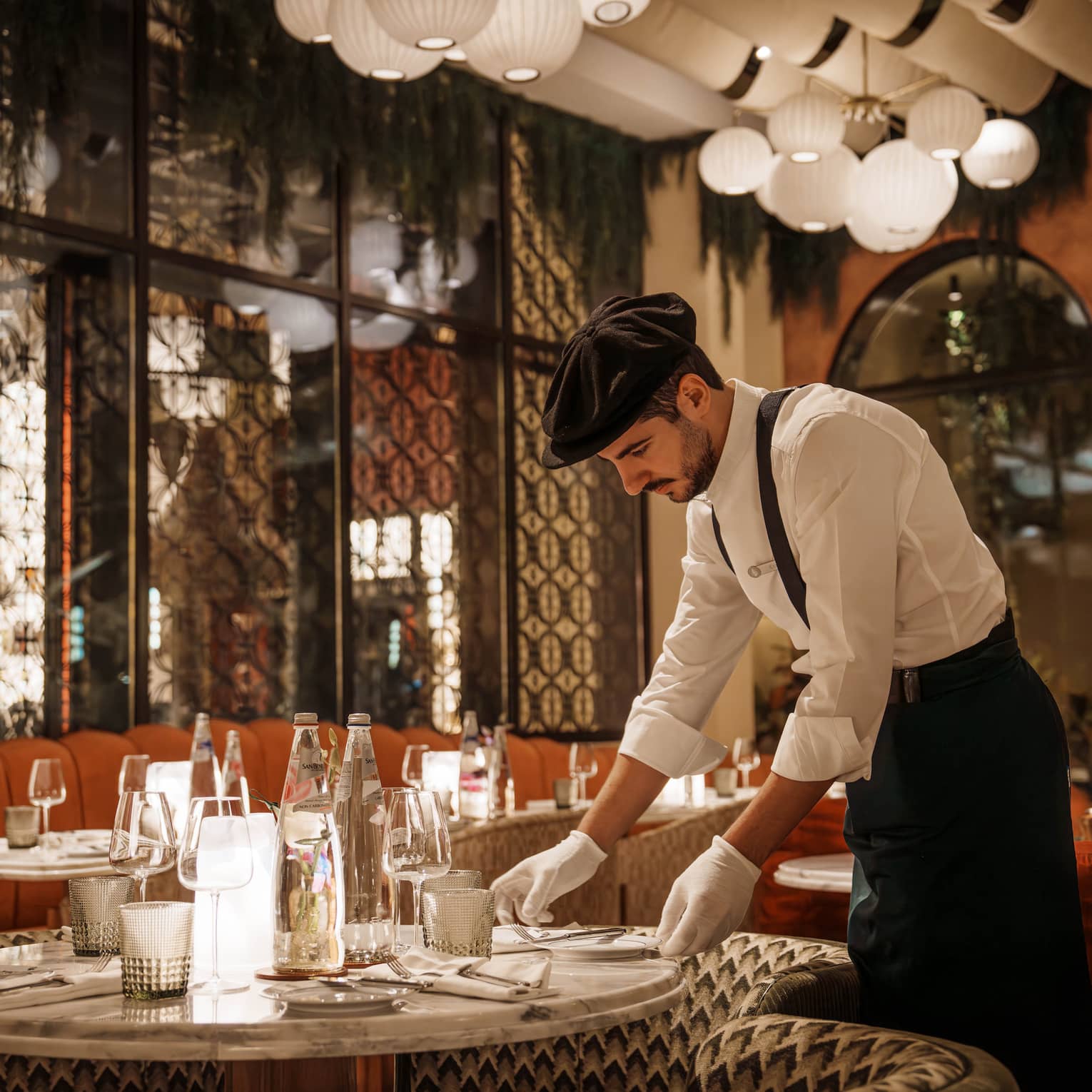 Waiter wearing white button-down shirt, black pants, black suspenders, black beret and white gloves arranges a plate on a table in an Art Deco?style restaurant