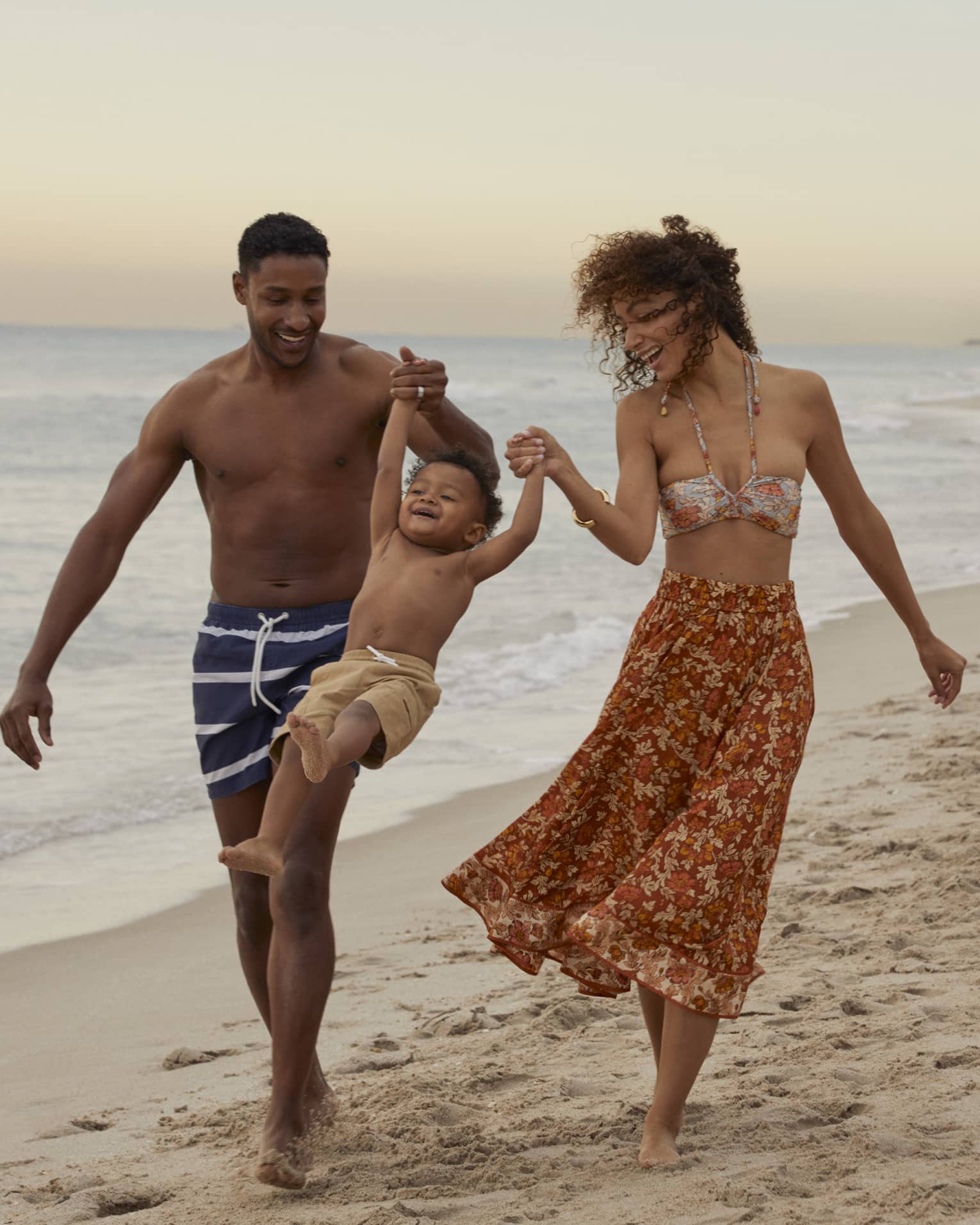 Smiling man and woman in beach attire swing young child by their arms as they walk on a Fort Lauderdale beach at sunset