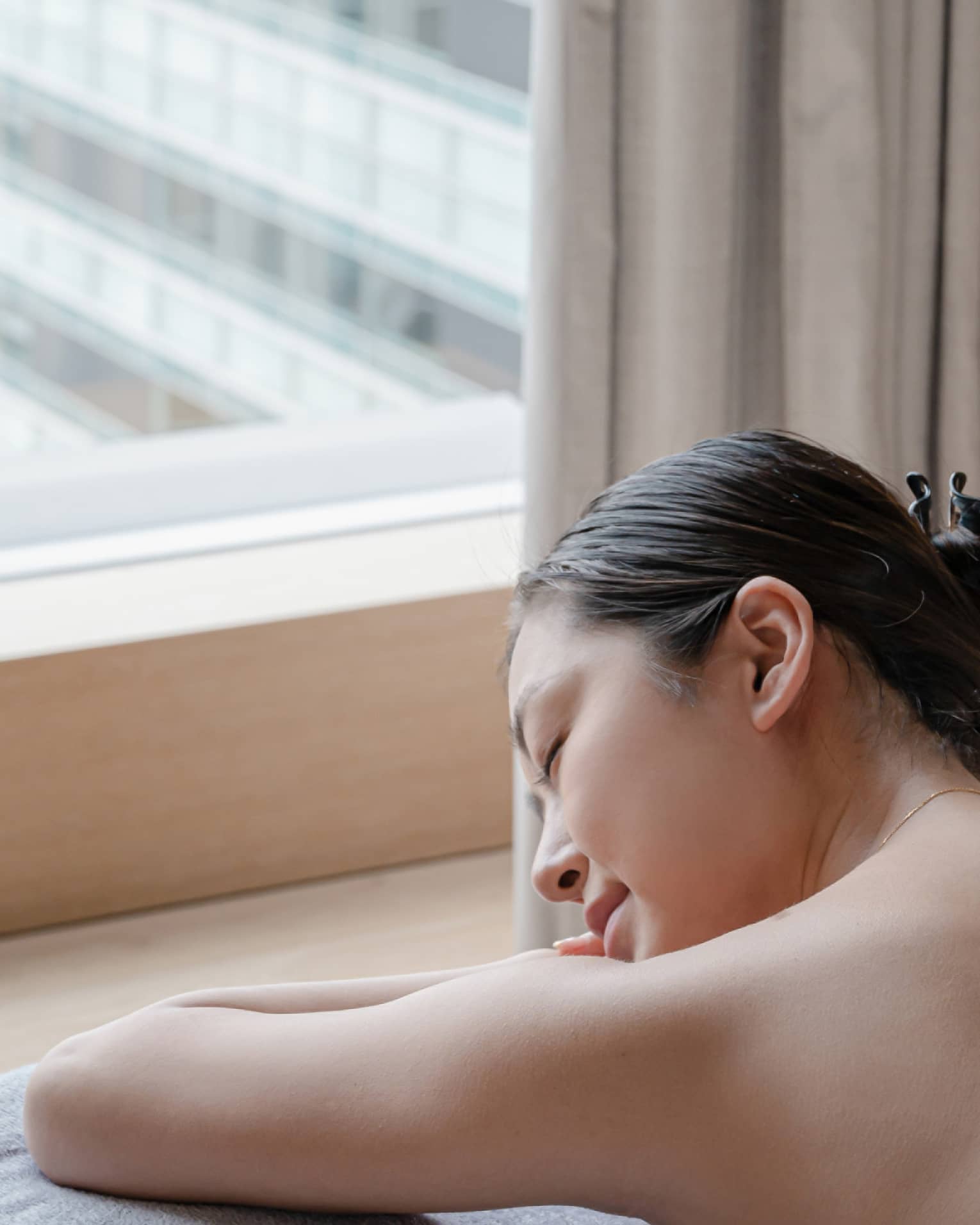 Person relaxing on massage table by a window with eyes closed, enjoying a peaceful moment