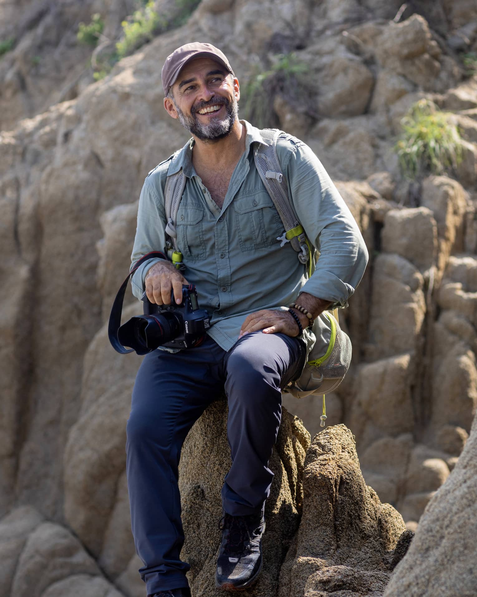 A photographer wearing a backpack and holding a camera in one hand sits atop a rock formation, smiling into the distance.