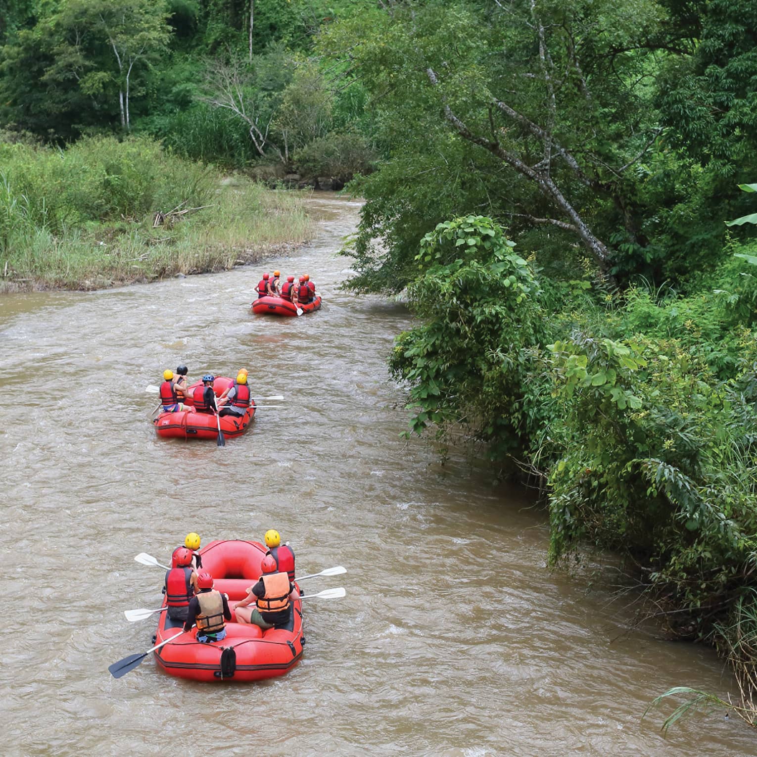 Rear view of three red inflatable rafts, five helmeted riders in each, coasting down a winding river flanked by lush foliage.