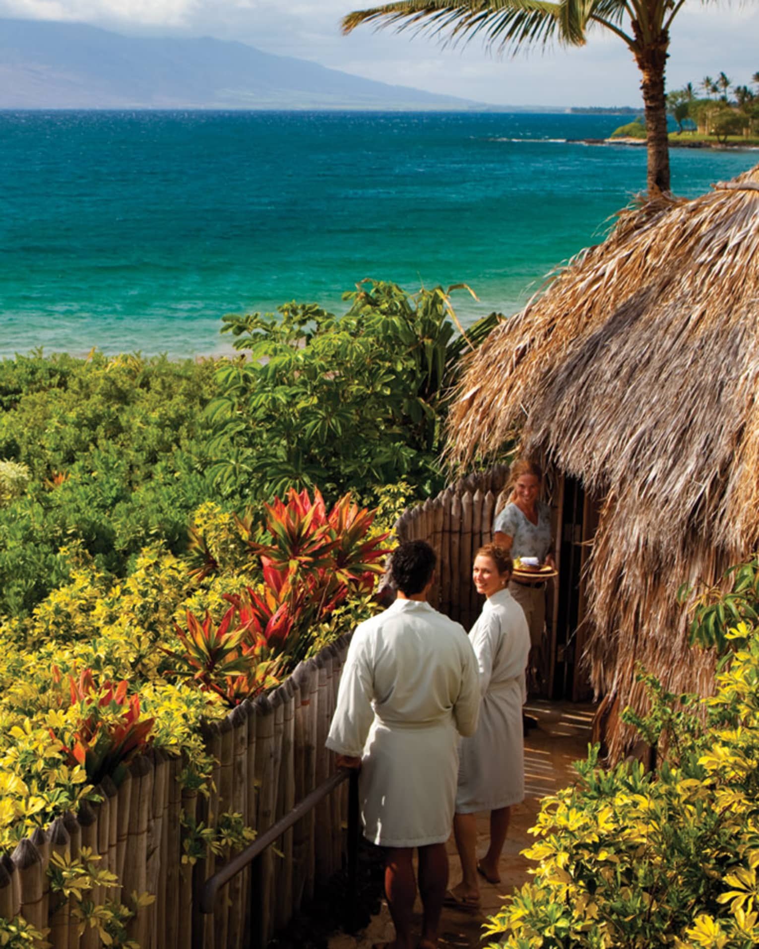 Couple in white bathrobes follow spa attendant into thatched-roof hut on path looking out at ocean