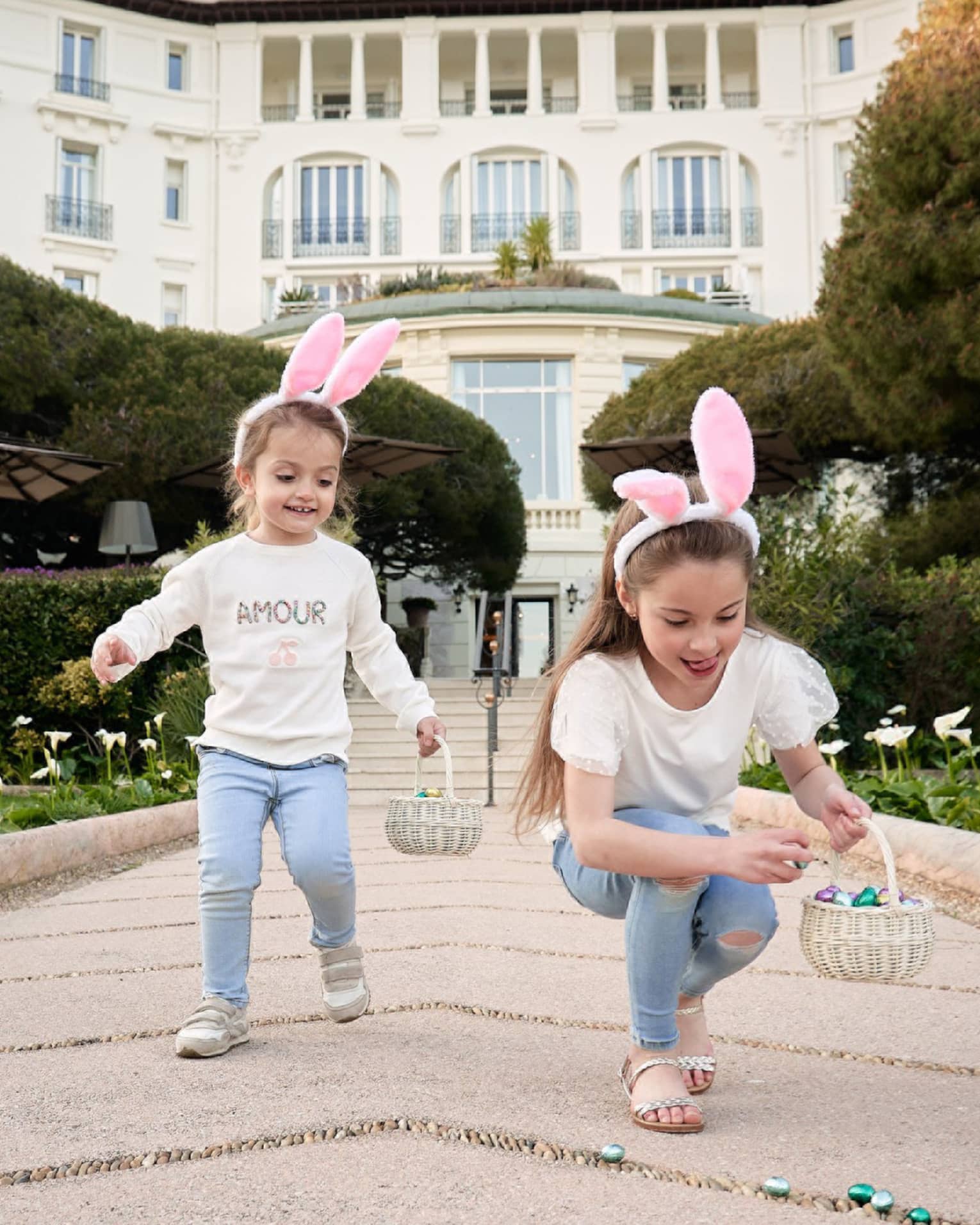 Two children in matching outfits wearing pink rabbit ears and holding Easter baskets with grand white building in backdrop