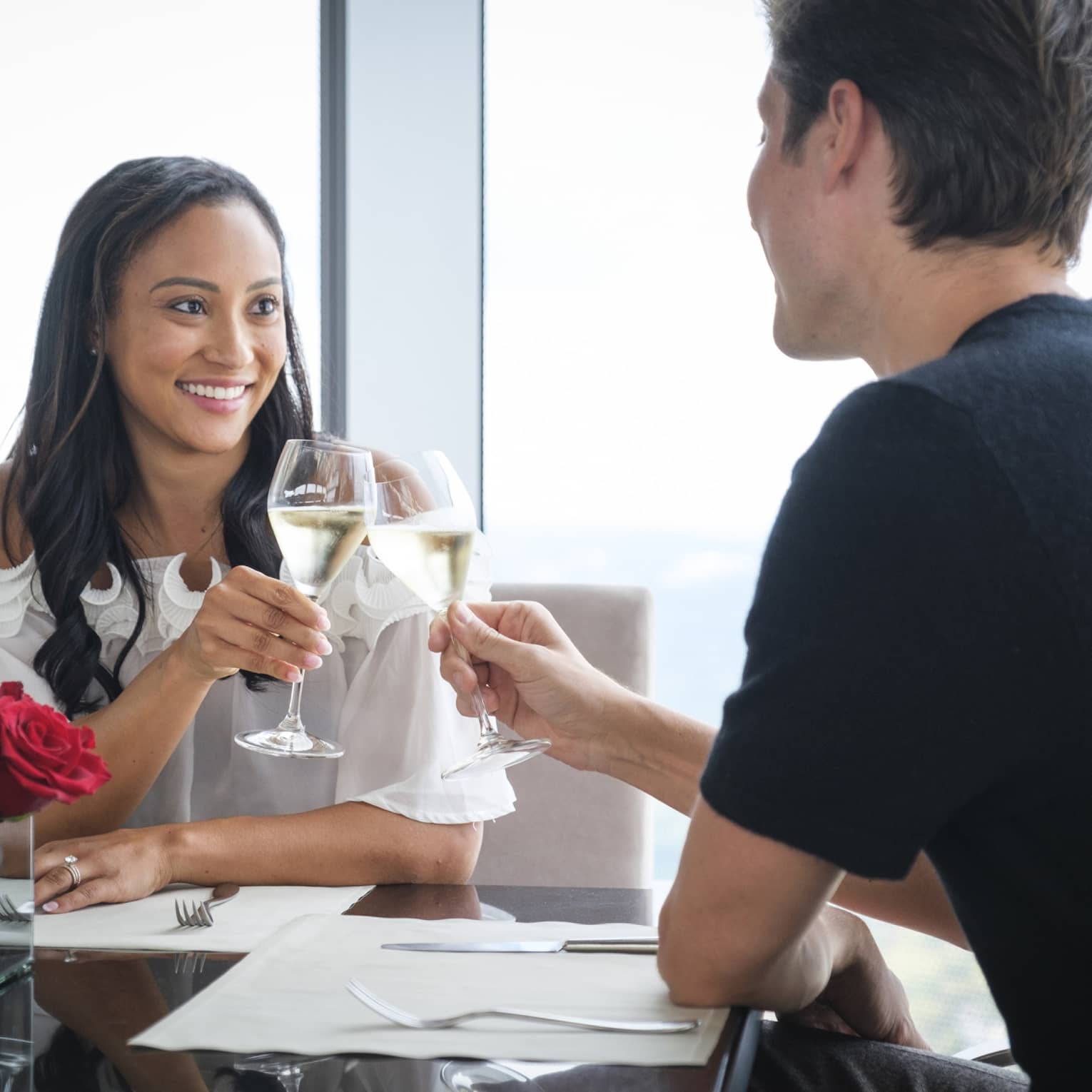 A man and woman celebrating with wine and red roses on a table near a large window.