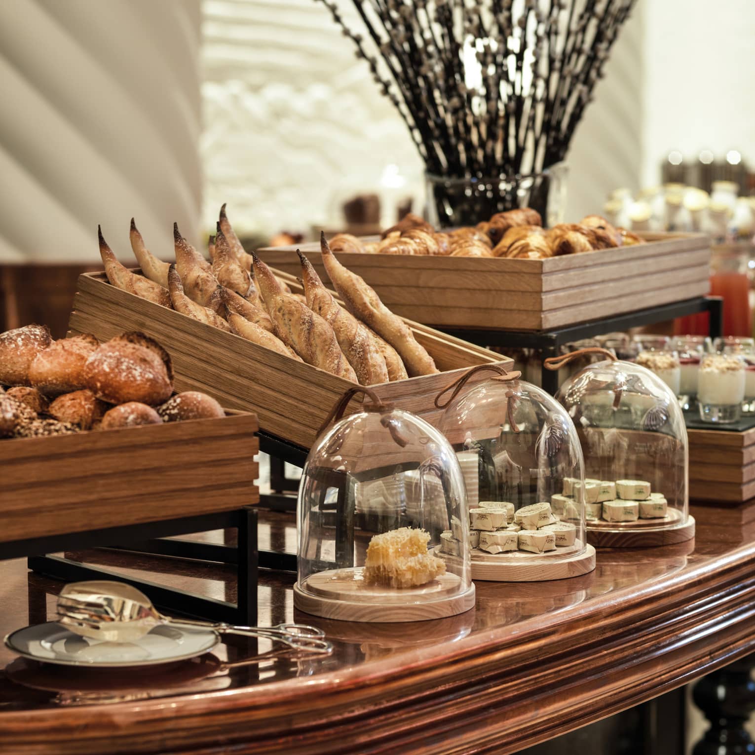 A decadent array of artisan breads and other baked goods in wooden crates and glass-covered dishes on a large wooden table.