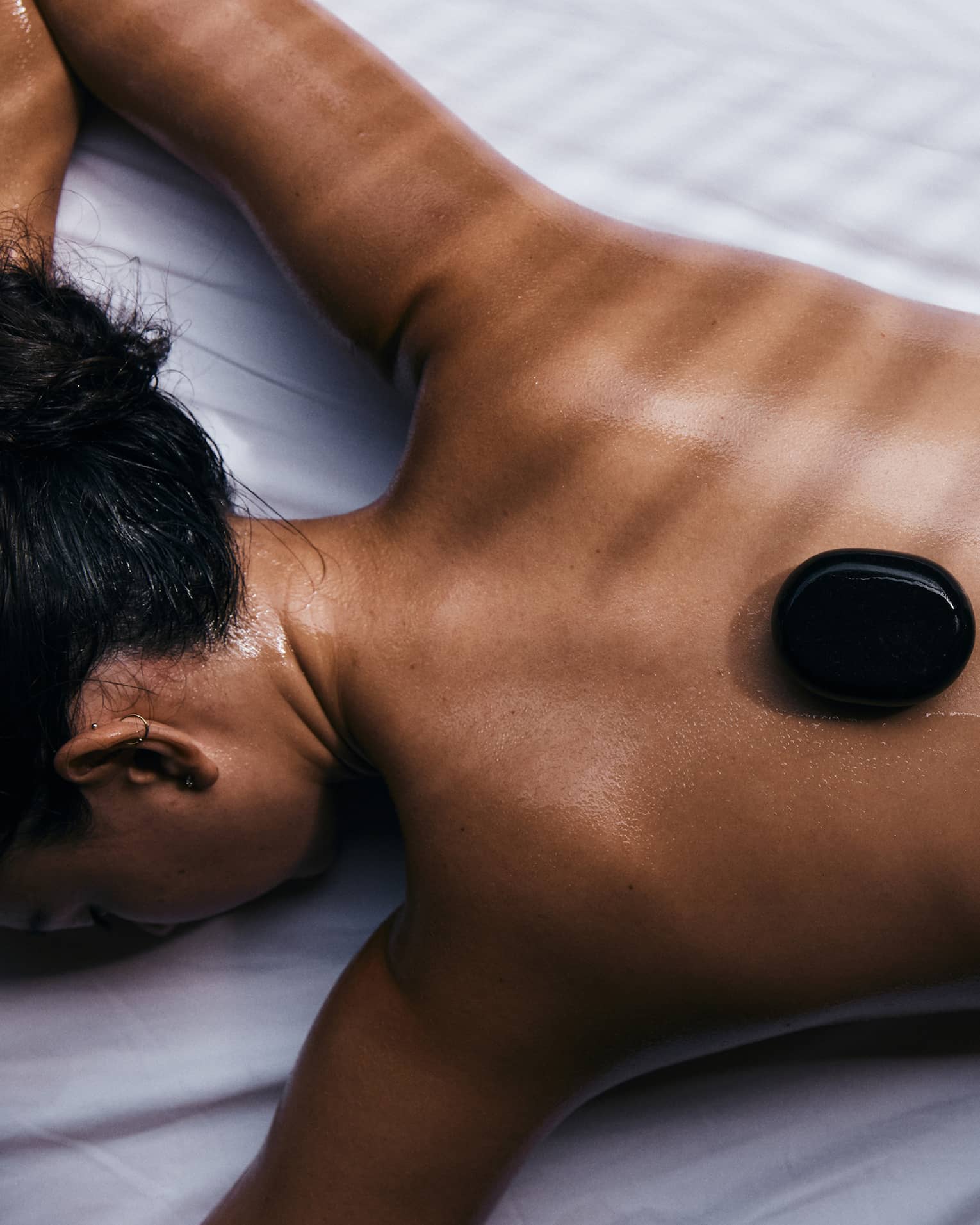 Close-up of a guest relaxing on their stomach on a bed of white linen with three black warming stones on their bare back.