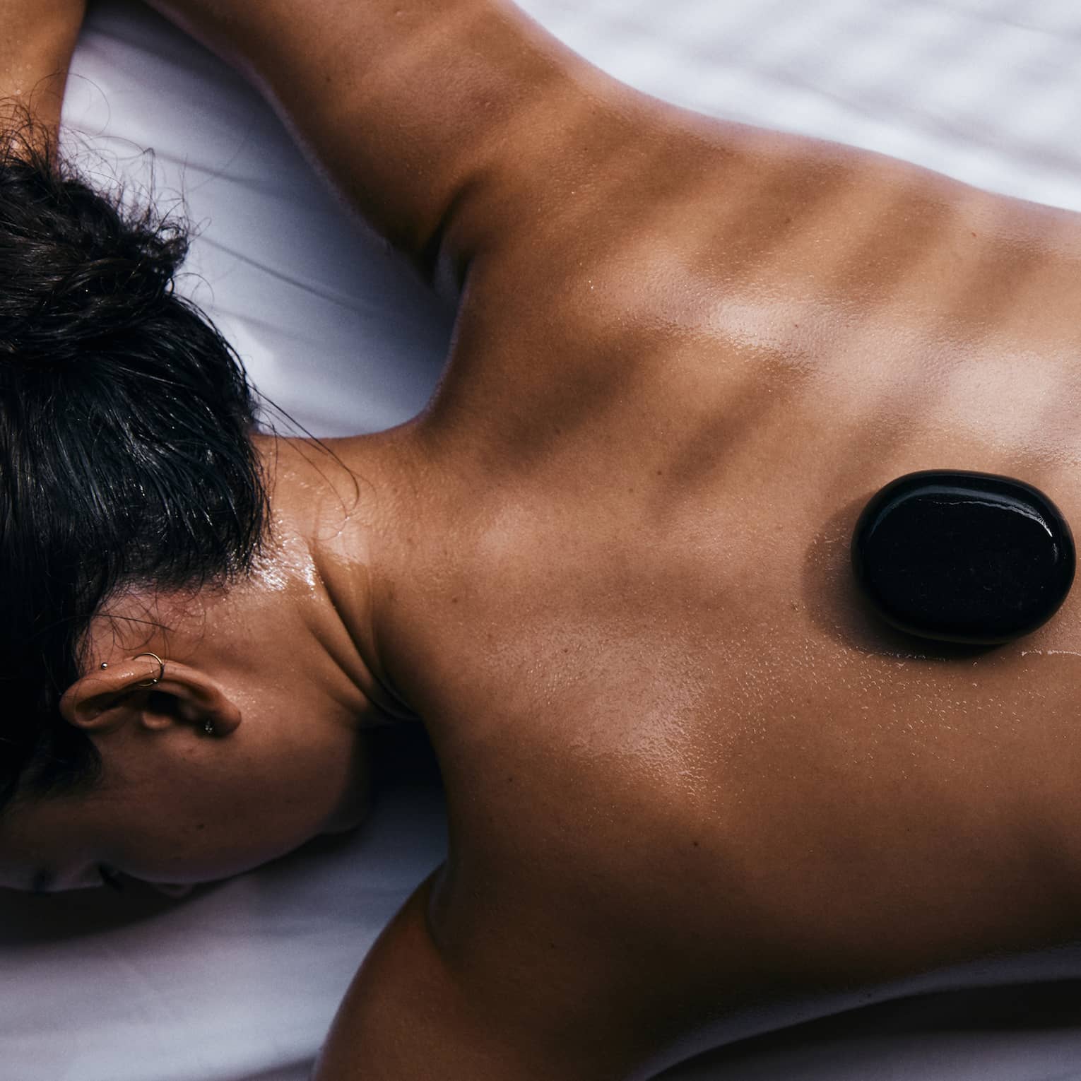 Close-up of a guest relaxing on their stomach on a bed of white linen with three black warming stones on their bare back.