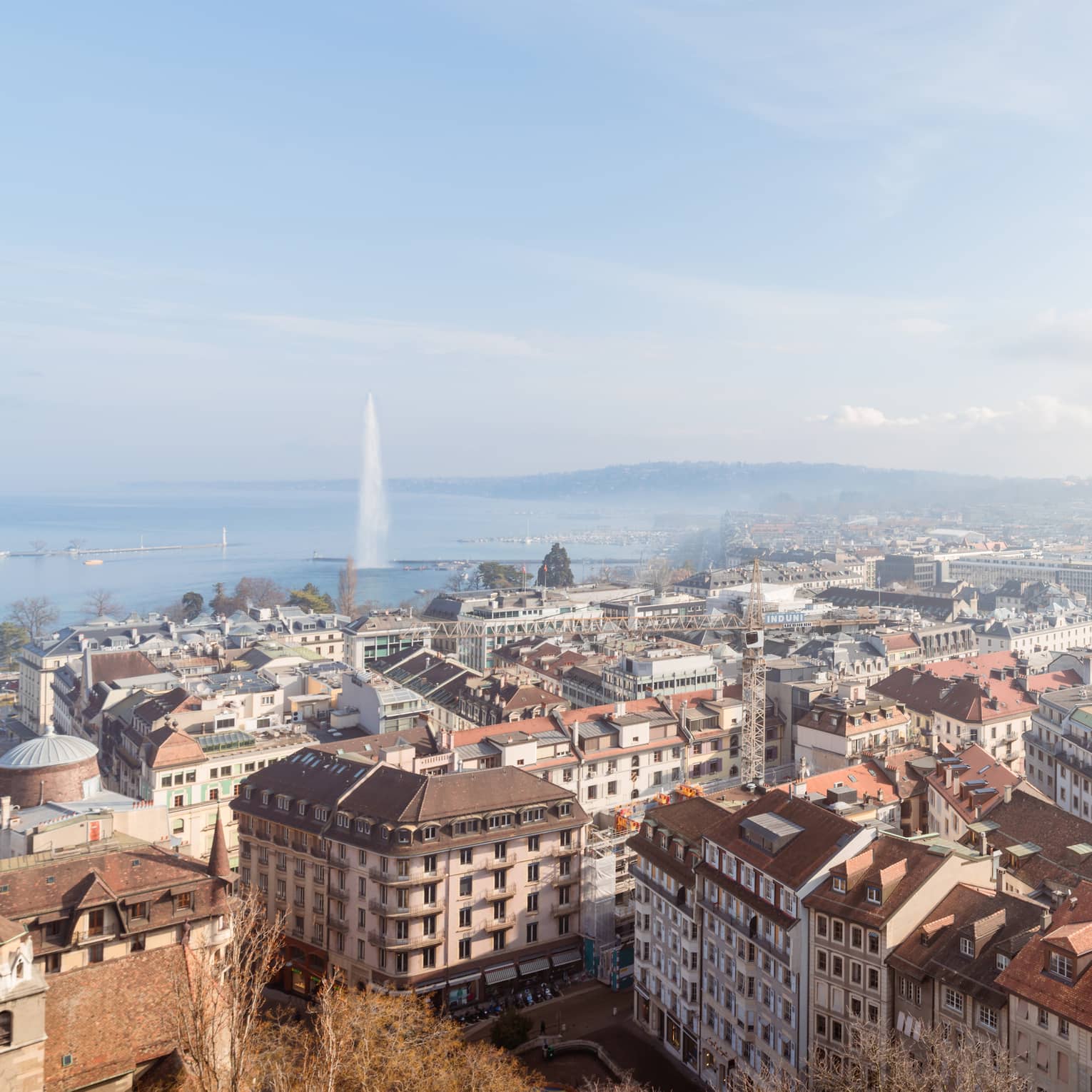 A cityscape with rooftops, buildings and streets, with a large fountain shooting water into the air over a lake in the background under a clear blue sky