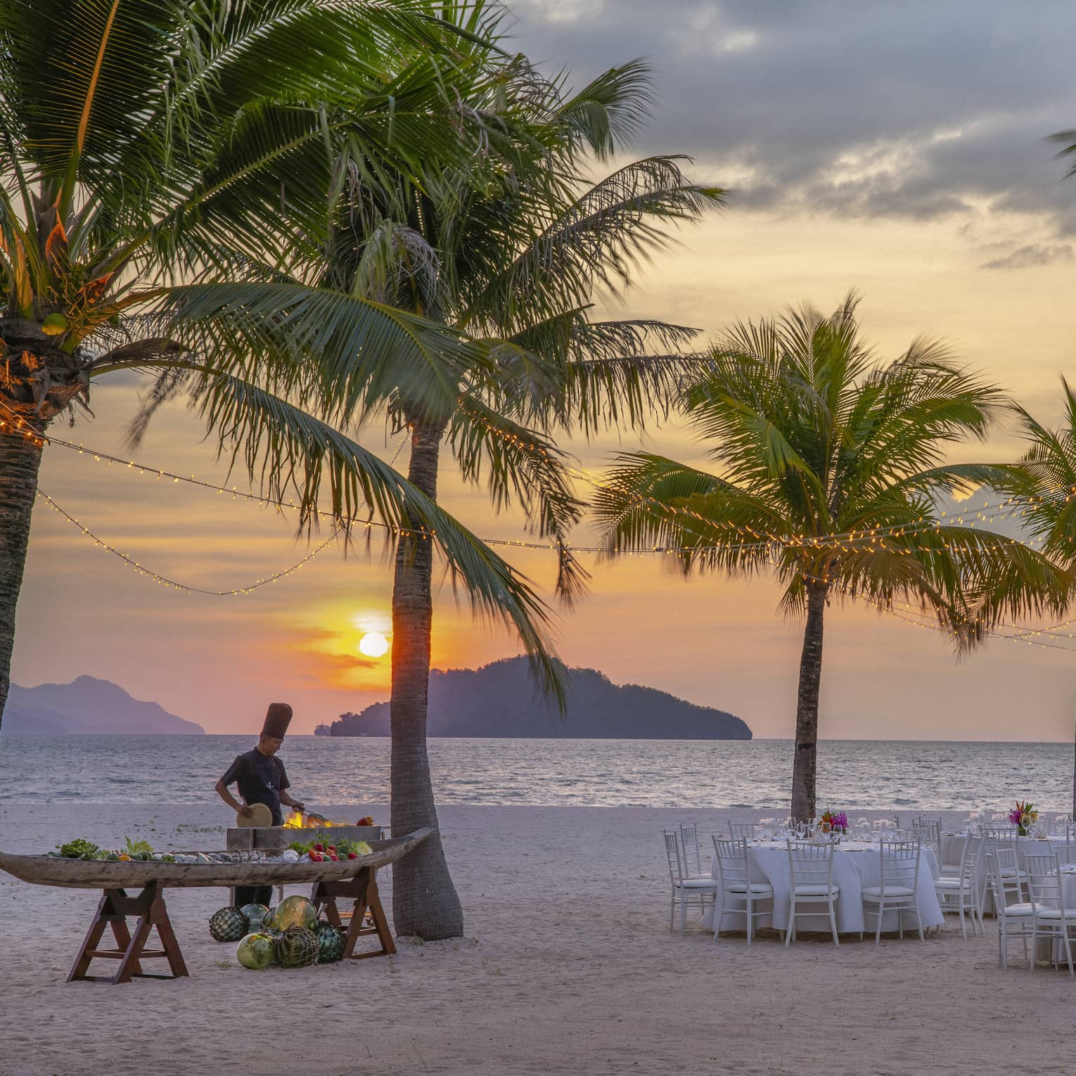 Tables set for dinner on the beach under a canopy of palm trees, a chef cooking over an open flame against the setting sun.