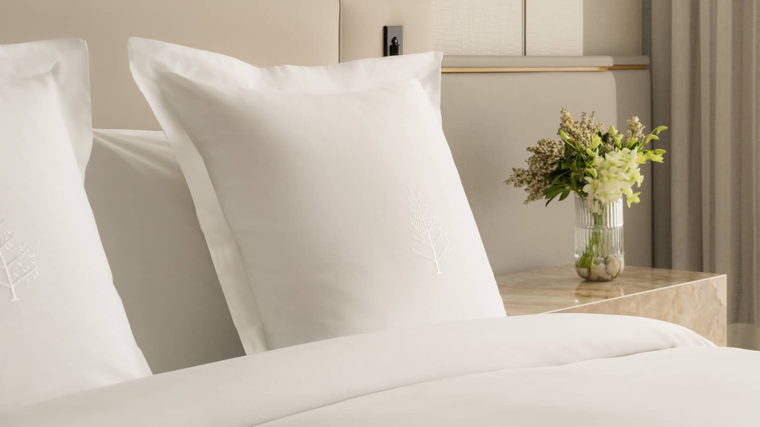Close-up of a hotel bed with white pillows embroidered with Four Seasons logos and a nightstand with a glass vase of fresh flowers
