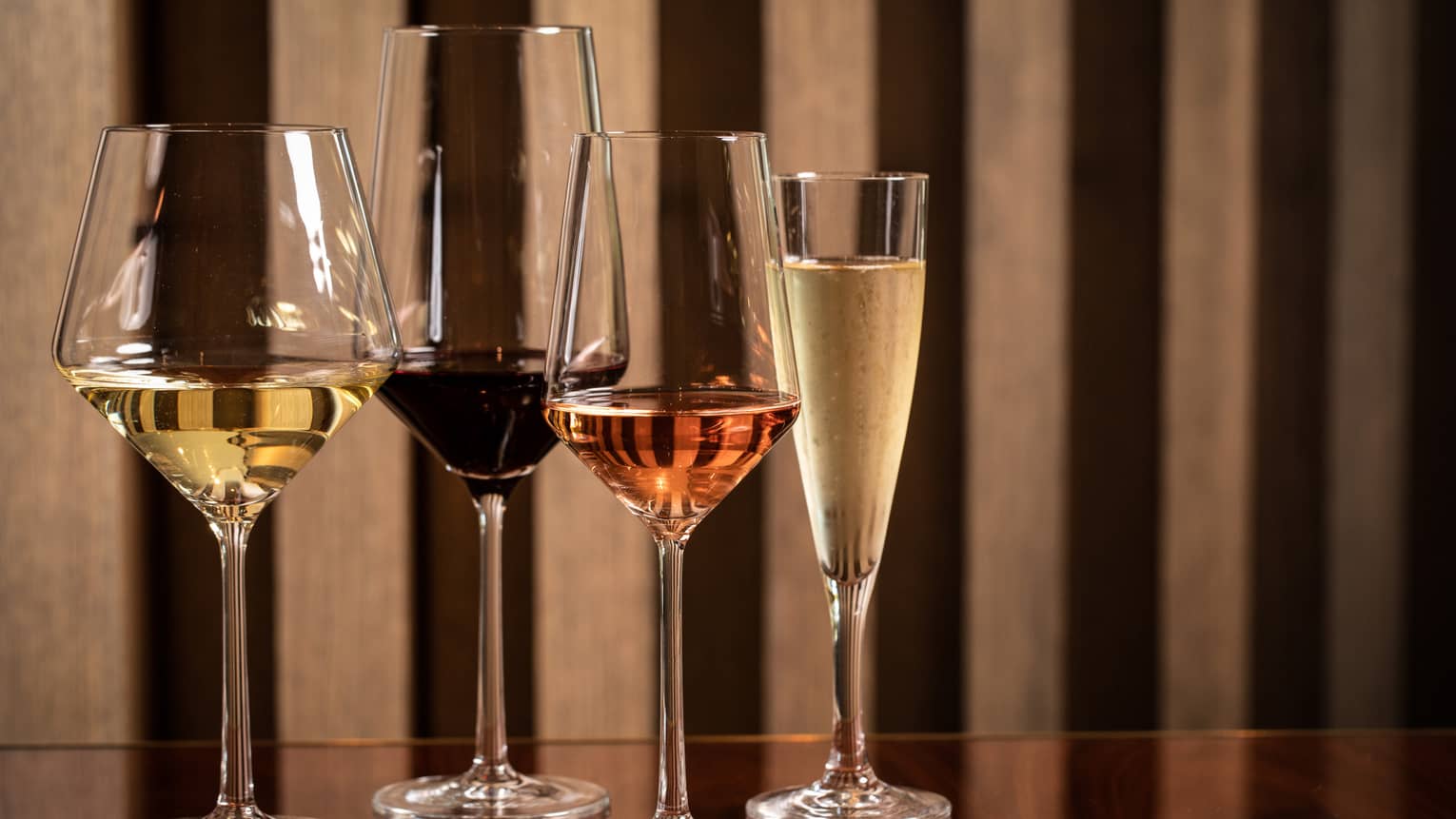 Four modern wine glasses holding white, red and rose wine are lined up on a wooden table 