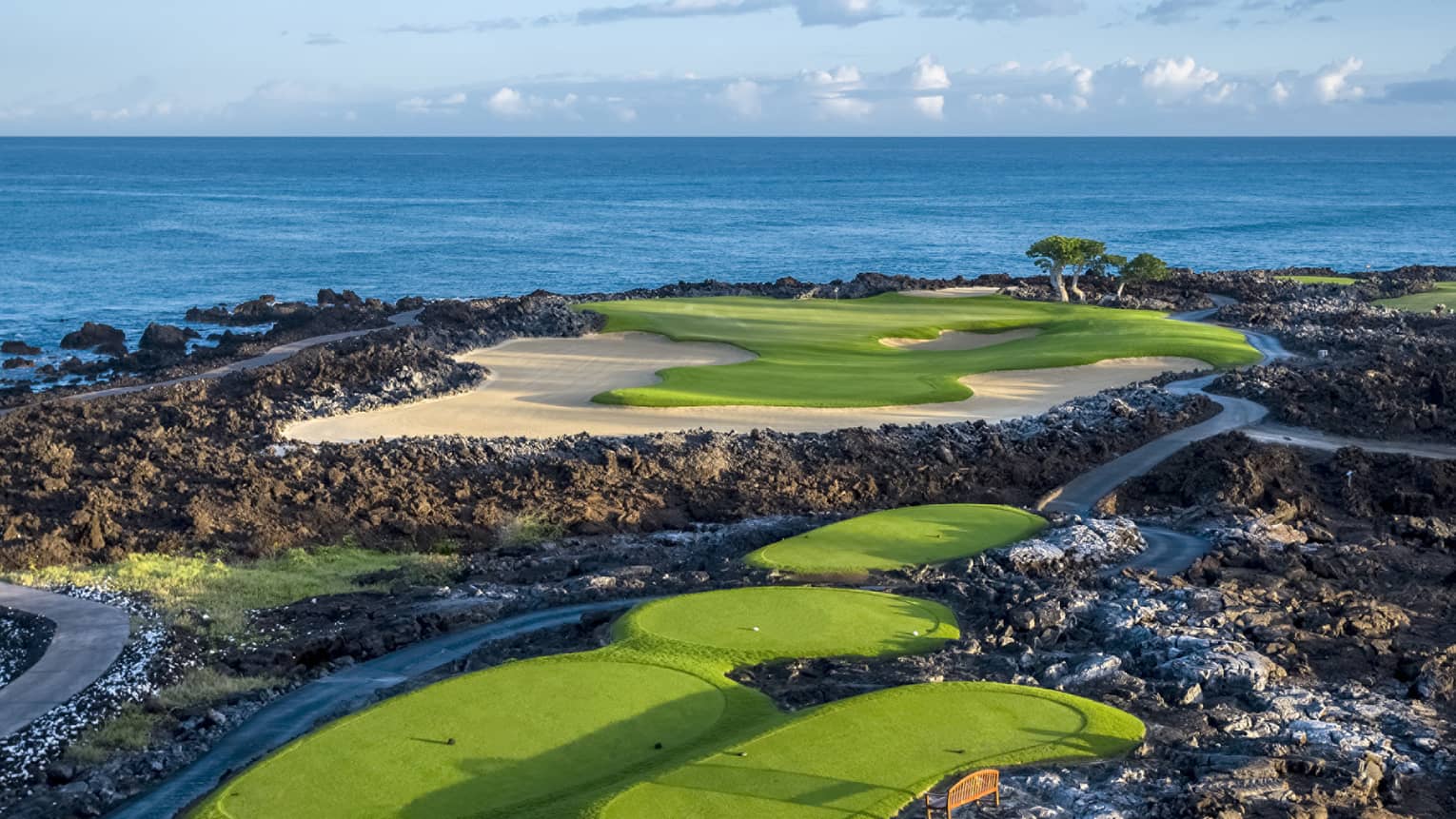 An impressive golf course overlooking the ocean, fairways and sand traps amid black lava formations, blue skies overhead.