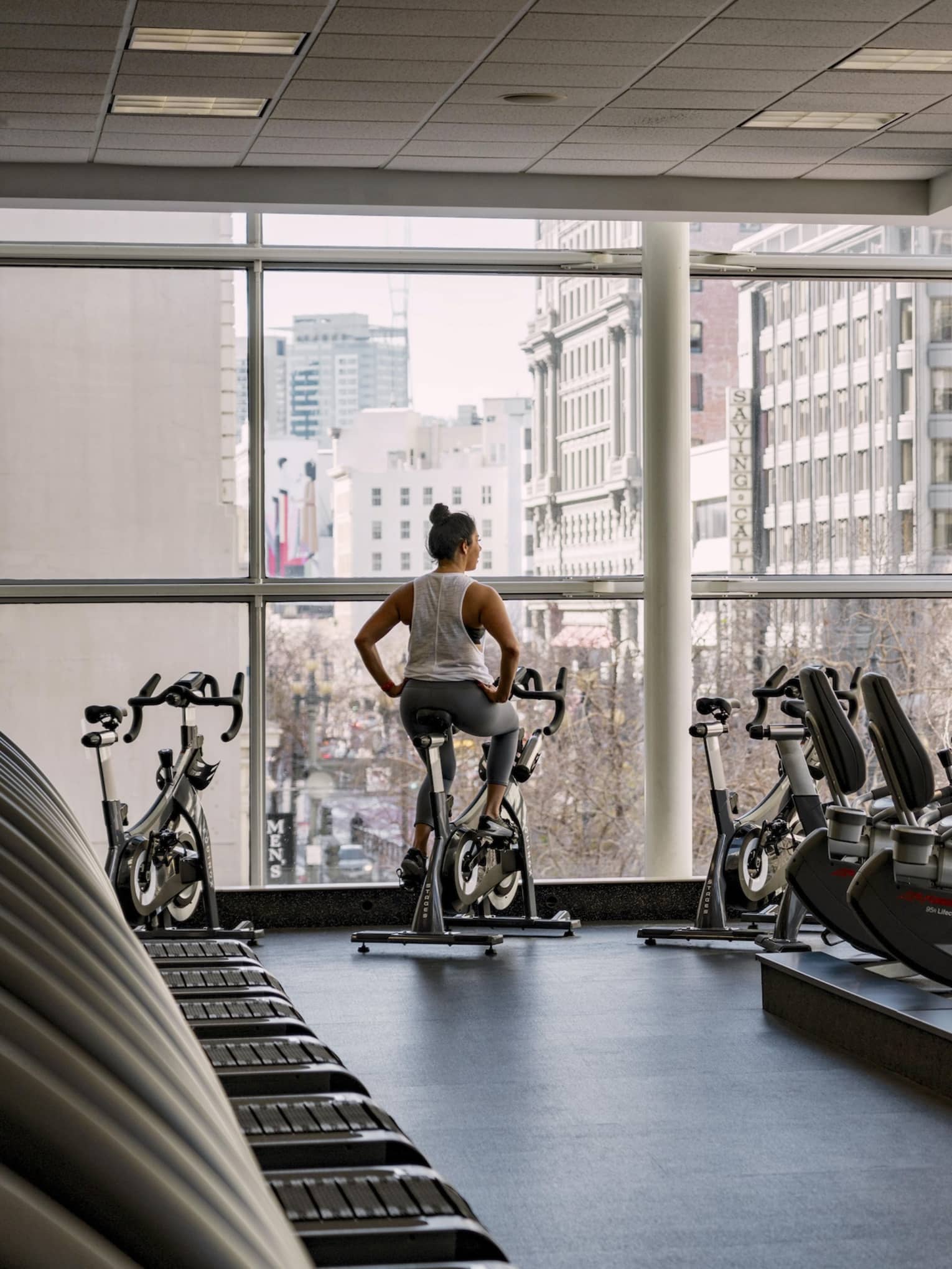 A woman rides a stationary bike in a hotel fitness center with floor-to-ceiling windows overlooking a city view