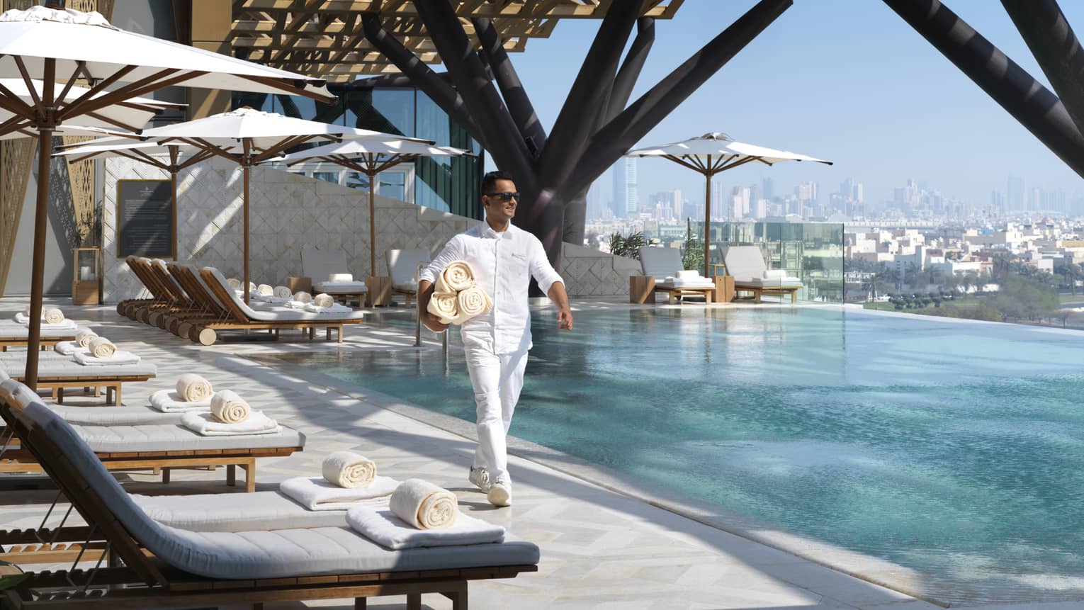 Hotel staff walks past white lounge chairs on outdoor swimming pool deck, carries rolled towels 
