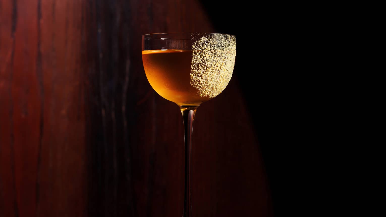 A tall stemmed red wine glass holds a golden-colored cocktail with gold dust sprinkled on the right side of the glass