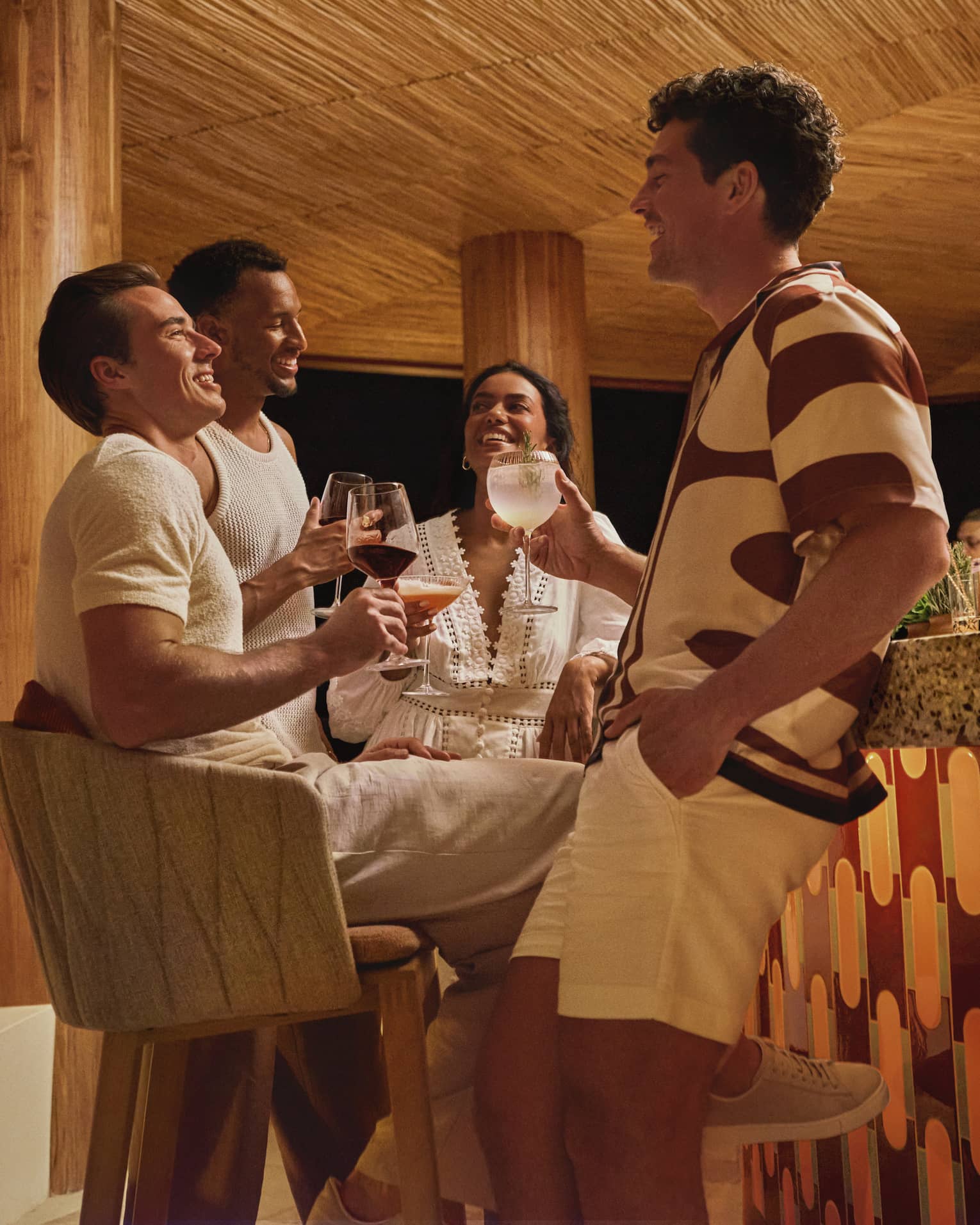 Four guests in stylish summer clothes hold cocktails and socialize next to a terazzo-topped bar in a round wooden pavilion.