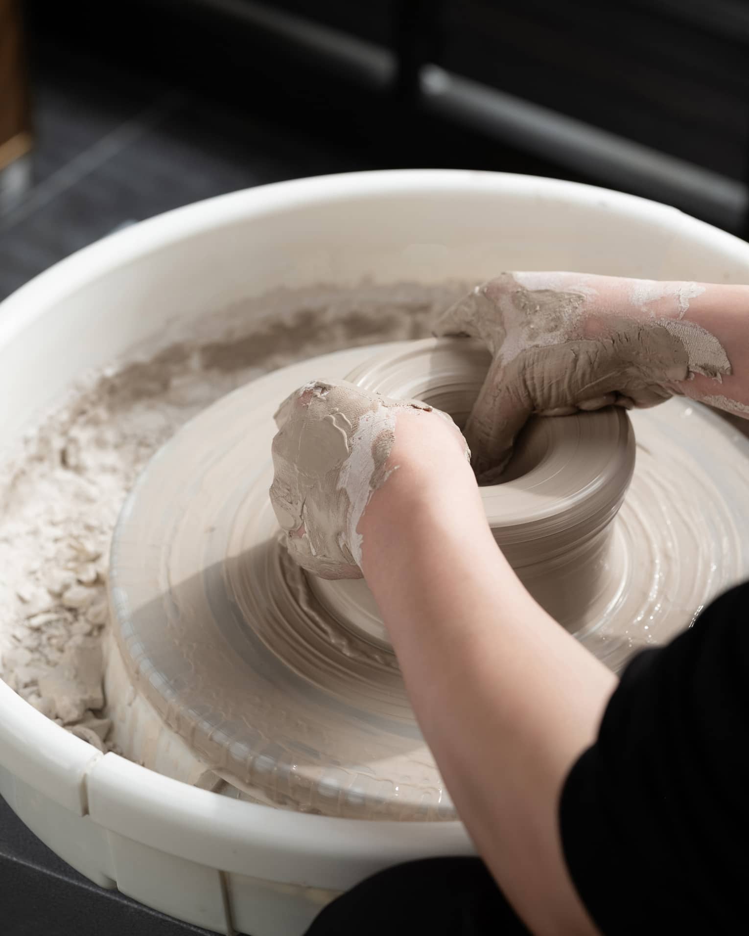Close-up of two hands covered in wet beige clay shaping a pot on a spinning pottery wheel. The potter is wearing black.
