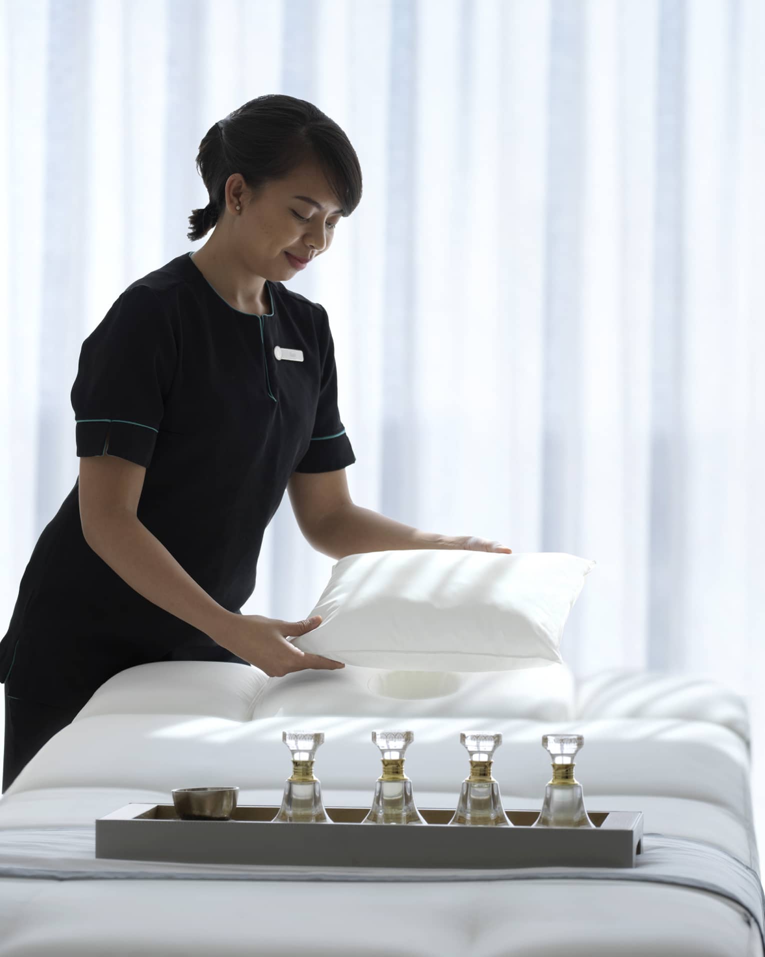 Spa attendant wearing a black uniform fluffs a while pillow on a treatment table covered with all-white linens and a tray filled with four small glass bottles