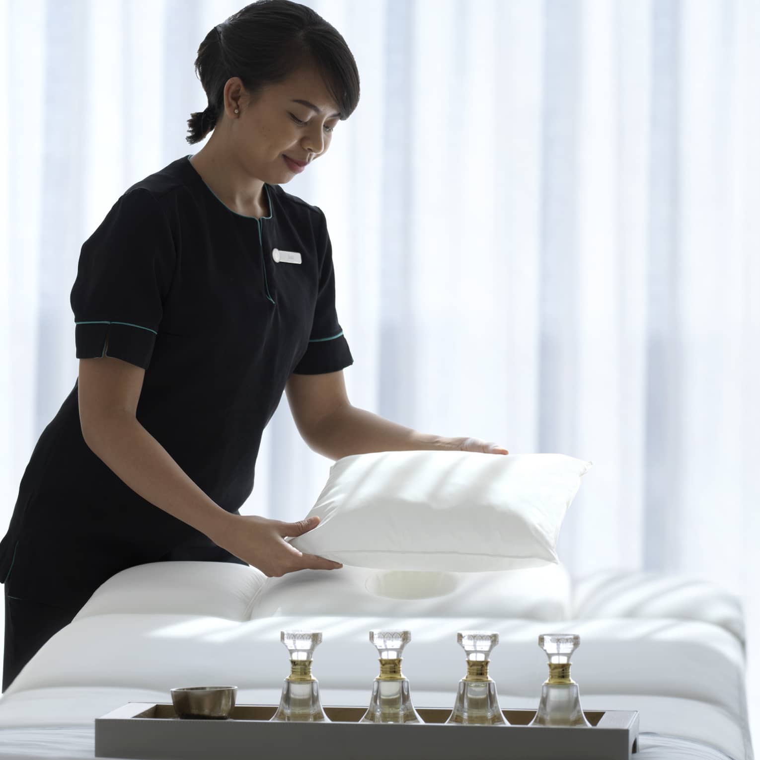 Spa attendant wearing a black uniform fluffs a while pillow on a treatment table covered with all-white linens and a tray filled with four small glass bottles