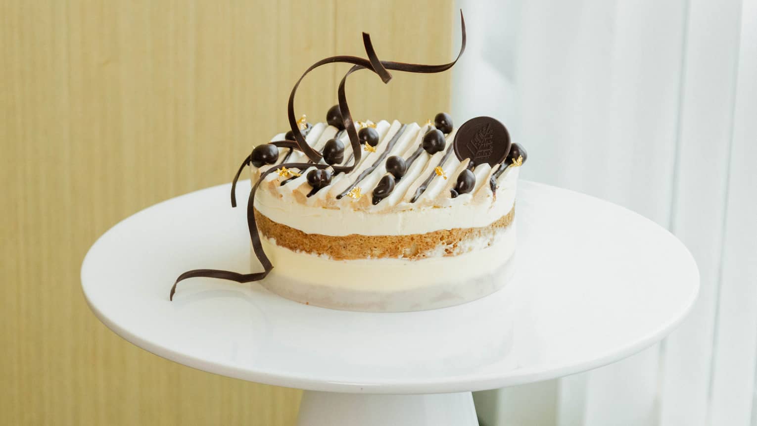 Round tiramisu topped with round chocolates and two long, thin chocolate twists is set atop a round white cake stand