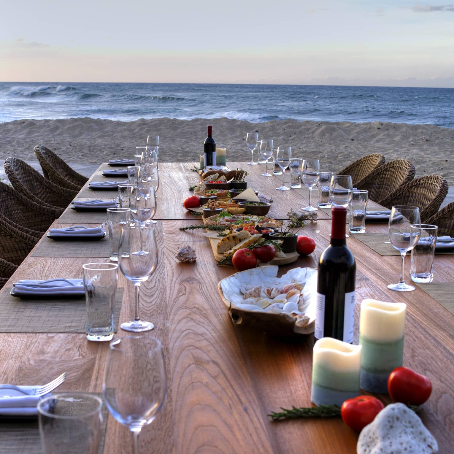 View down long wood private dining tale with wine, candles and shells, wicker chairs on beach