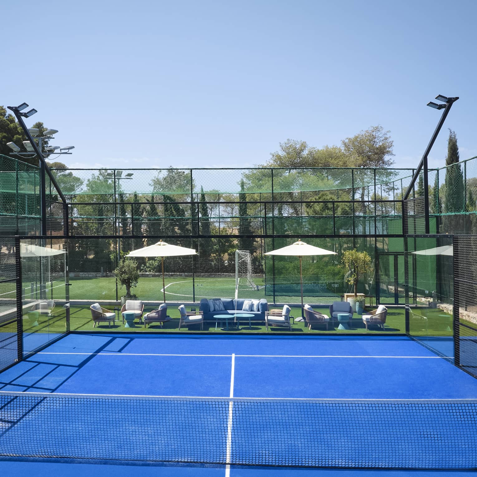 Bright blue padel court surrounded by fencing, white umbrellas and lounge chairs