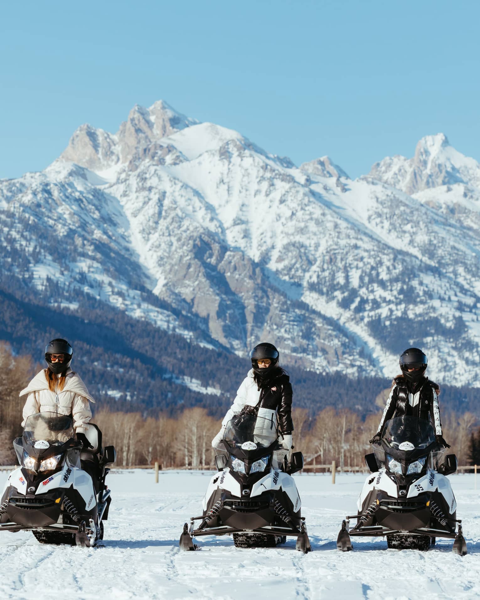 Three snowmobilers in stylish gear stand side-by-side on their machines facing the camera; a big snowy mountain looms behind.