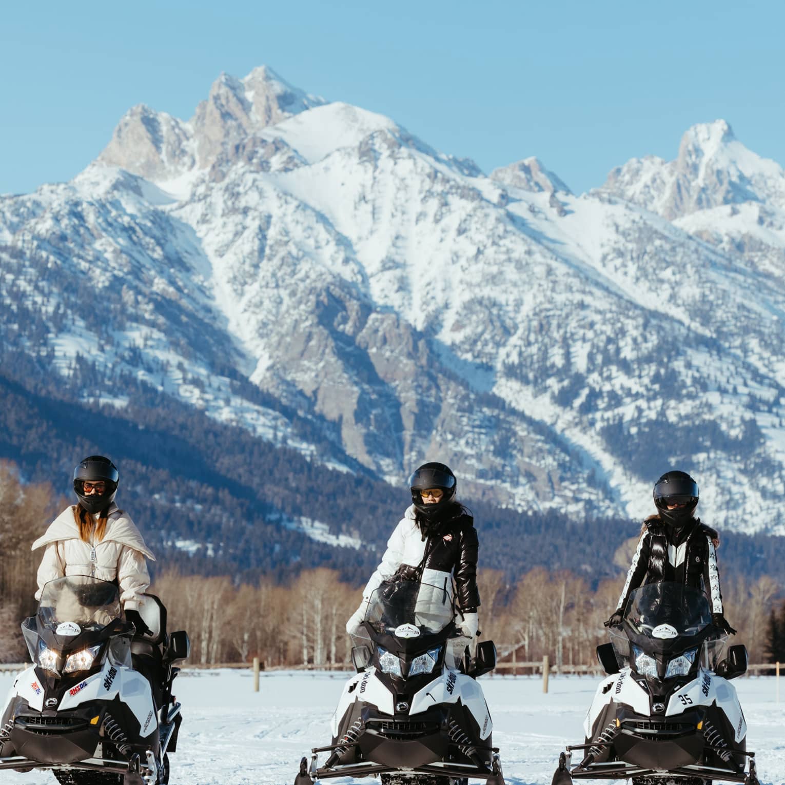 Three snowmobilers in stylish gear stand side-by-side on their machines facing the camera; a big snowy mountain looms behind.