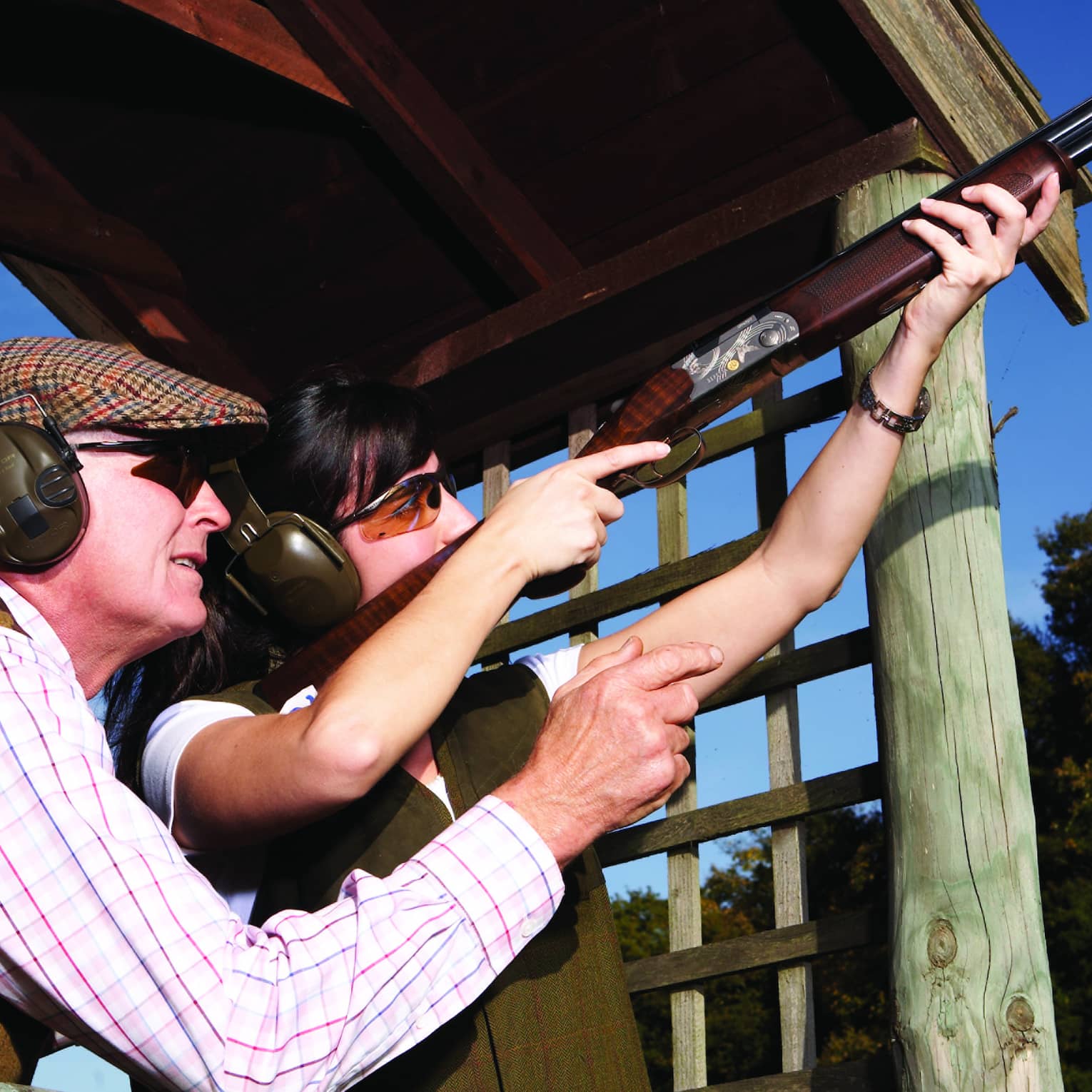 A close-up of two guests wearing tweed vests and shooting ear muffs aiming a rifle towards the clear blue sky.