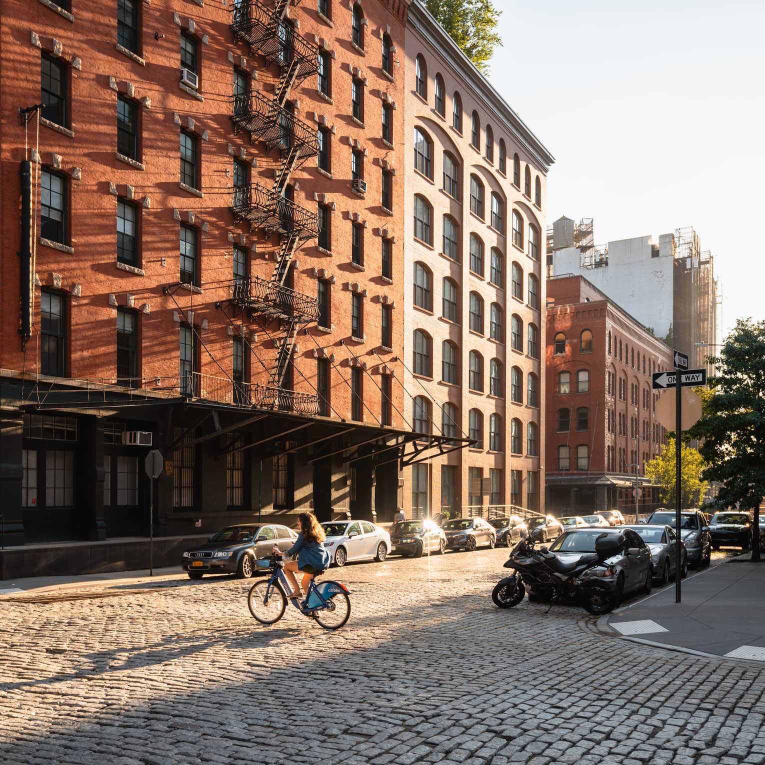 A person riding a bicycle in front of of a brick building with cars lining the street.