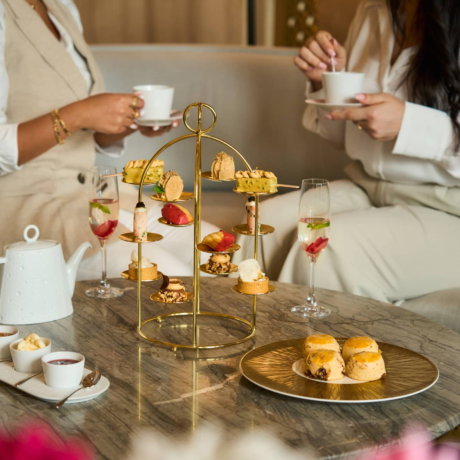 Two people sit on a beige sofa next to a marble coffee table set with a golden tray holding a variety of pastries, a white tea pot and othe small plates of food