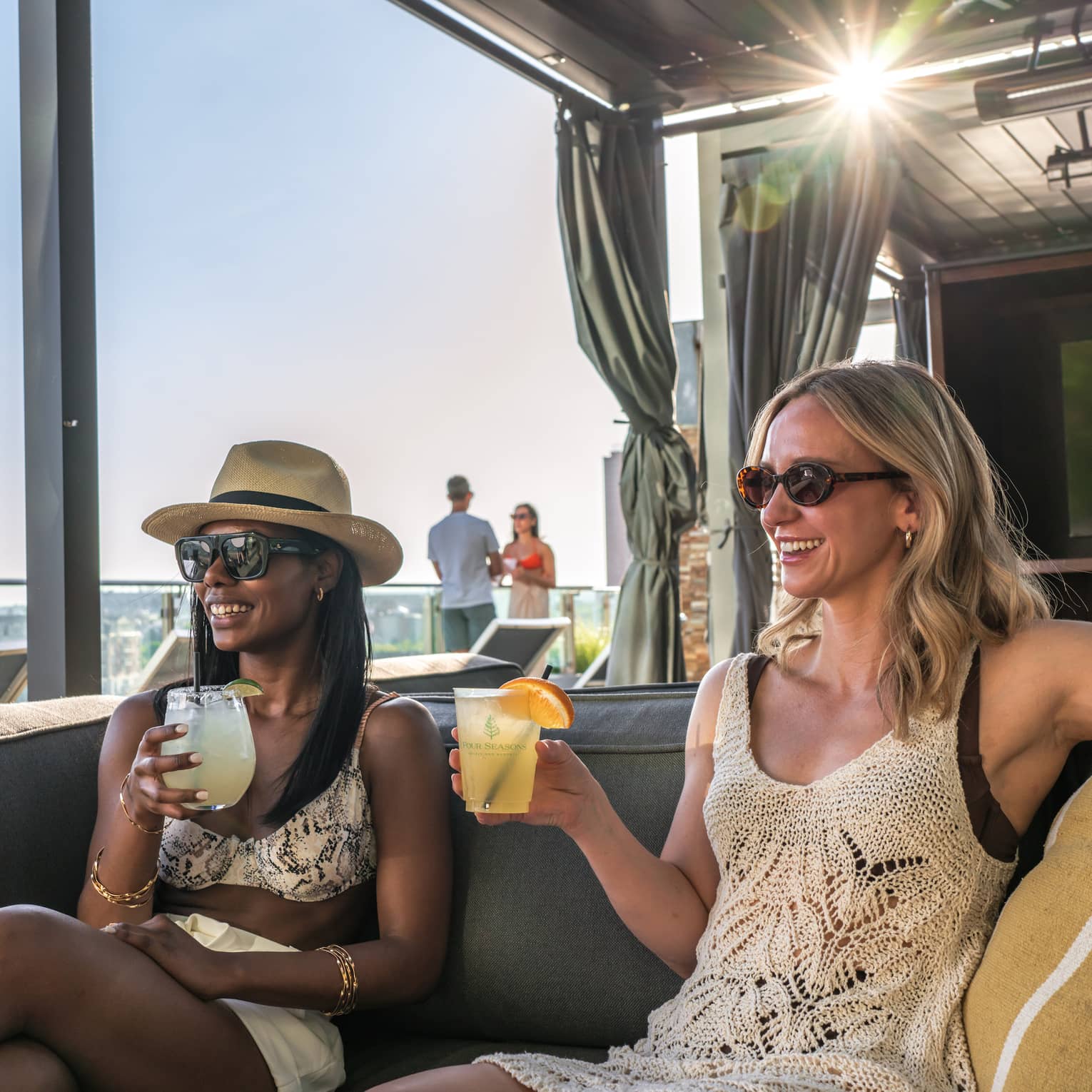 Two smiling friends holding iced beverages lounge on a cabana sofa, a bright burst of sunlight streaming through behind them.