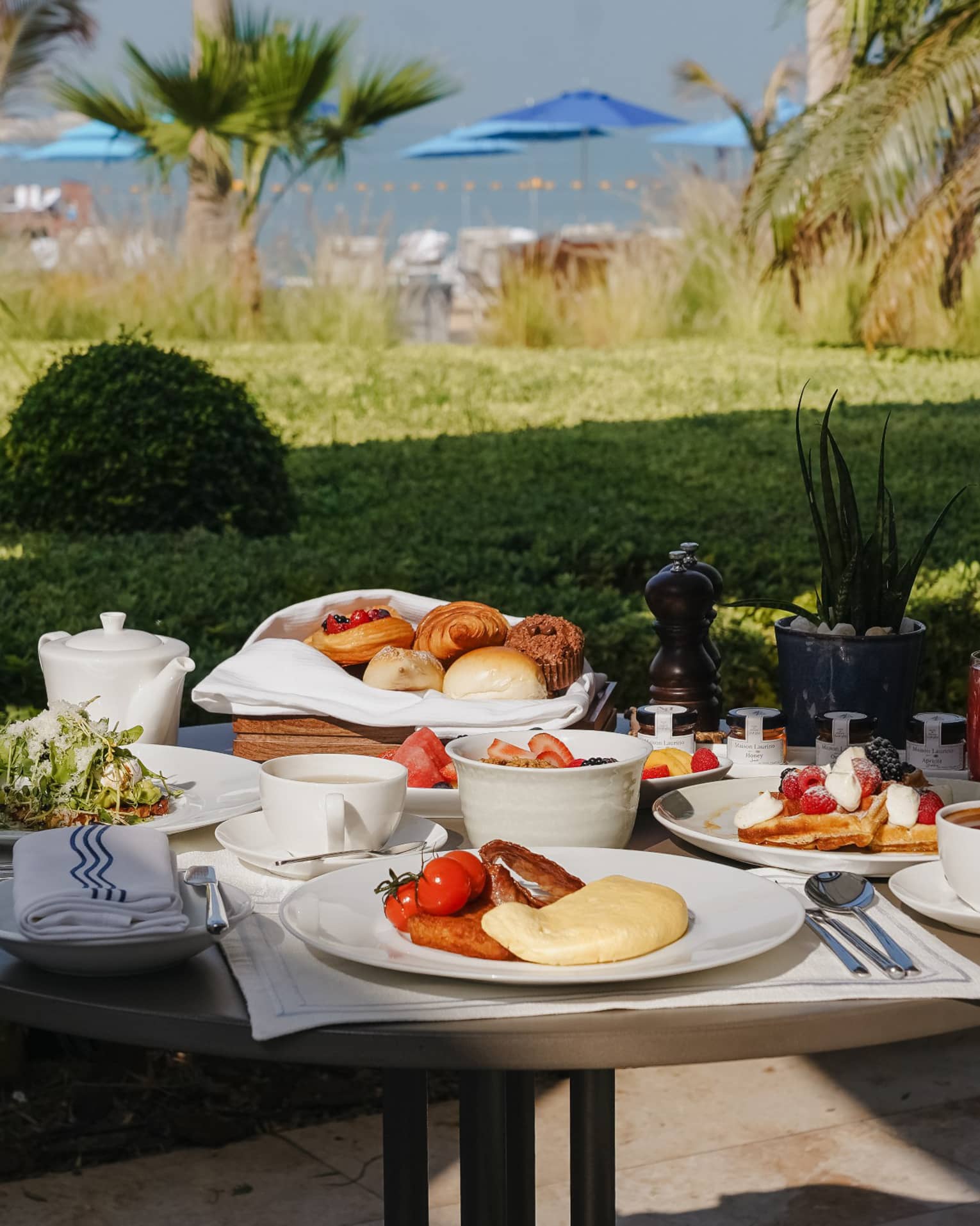 Outoor table filled with plates and bowls of breakfast food with a green lawn nearby and the ocean in the distance