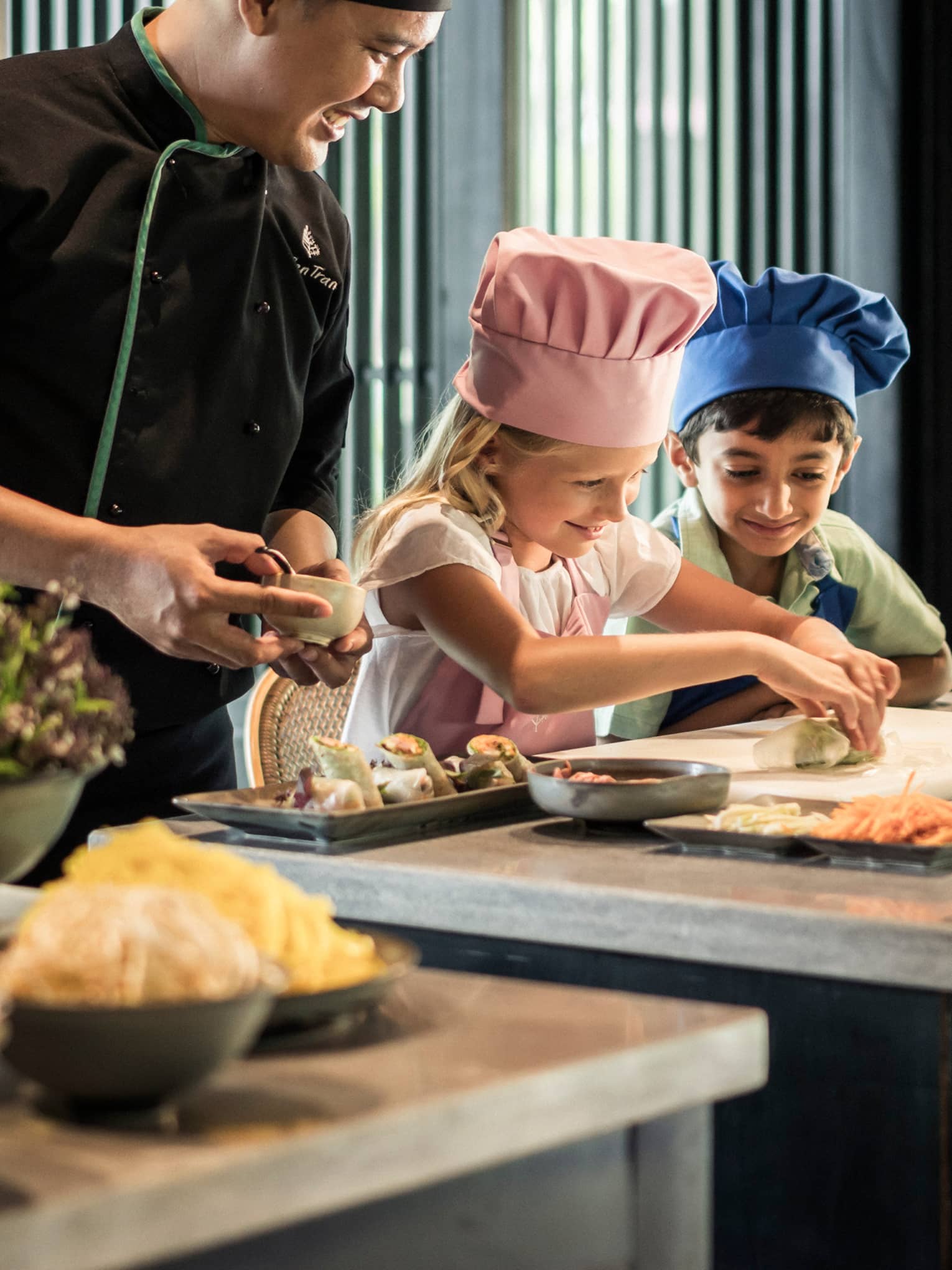 Chef watches as children wearing tall chef hats make spring rolls at counter