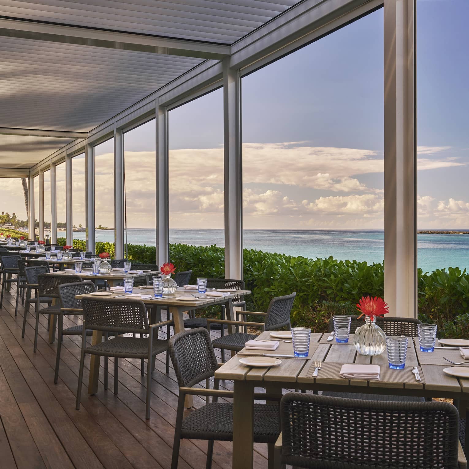 An outdoor eating area with square wood tables next to plants and the ocean.
