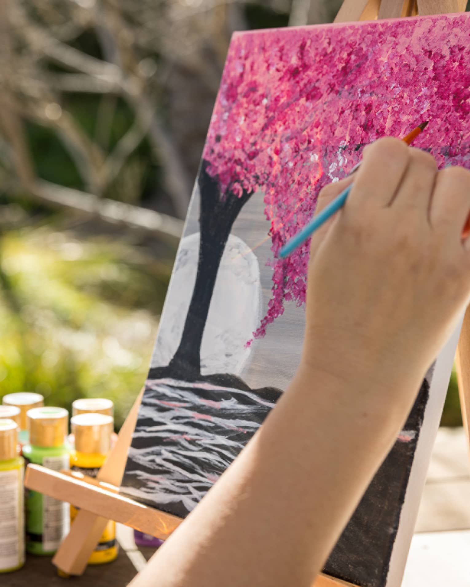 Person painting a pink tree on a canvas with one hand while holding a glass of wine in their other hand