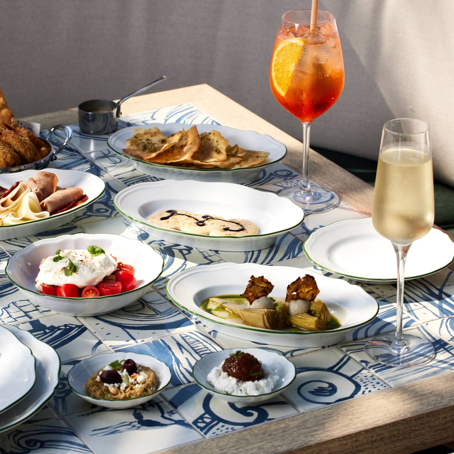 Outdoor table filled with a variety of food on white plates and three stemmed glasses filled with wine and cocktails