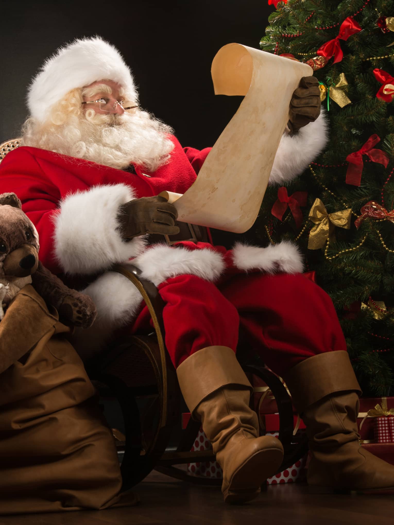 Santa Claus seated in large rocker holding a scroll, flanked by decorated Christmas tree with gifts and overflowing gift bag
