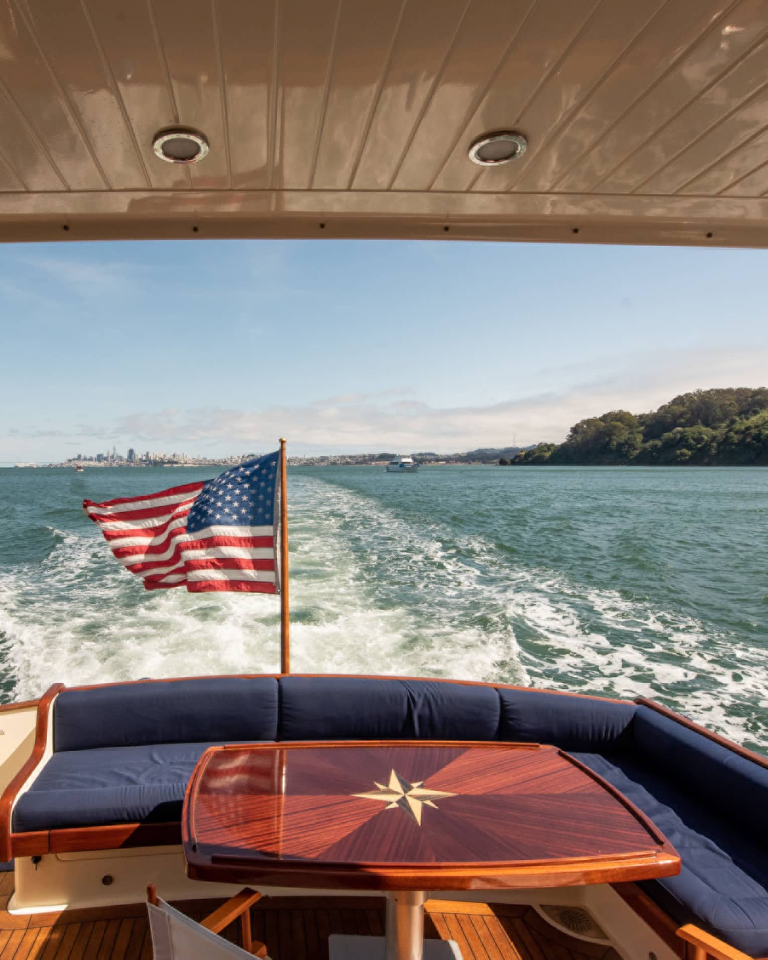 The back of a yacht, with a red table and an American flag, moving along the water.