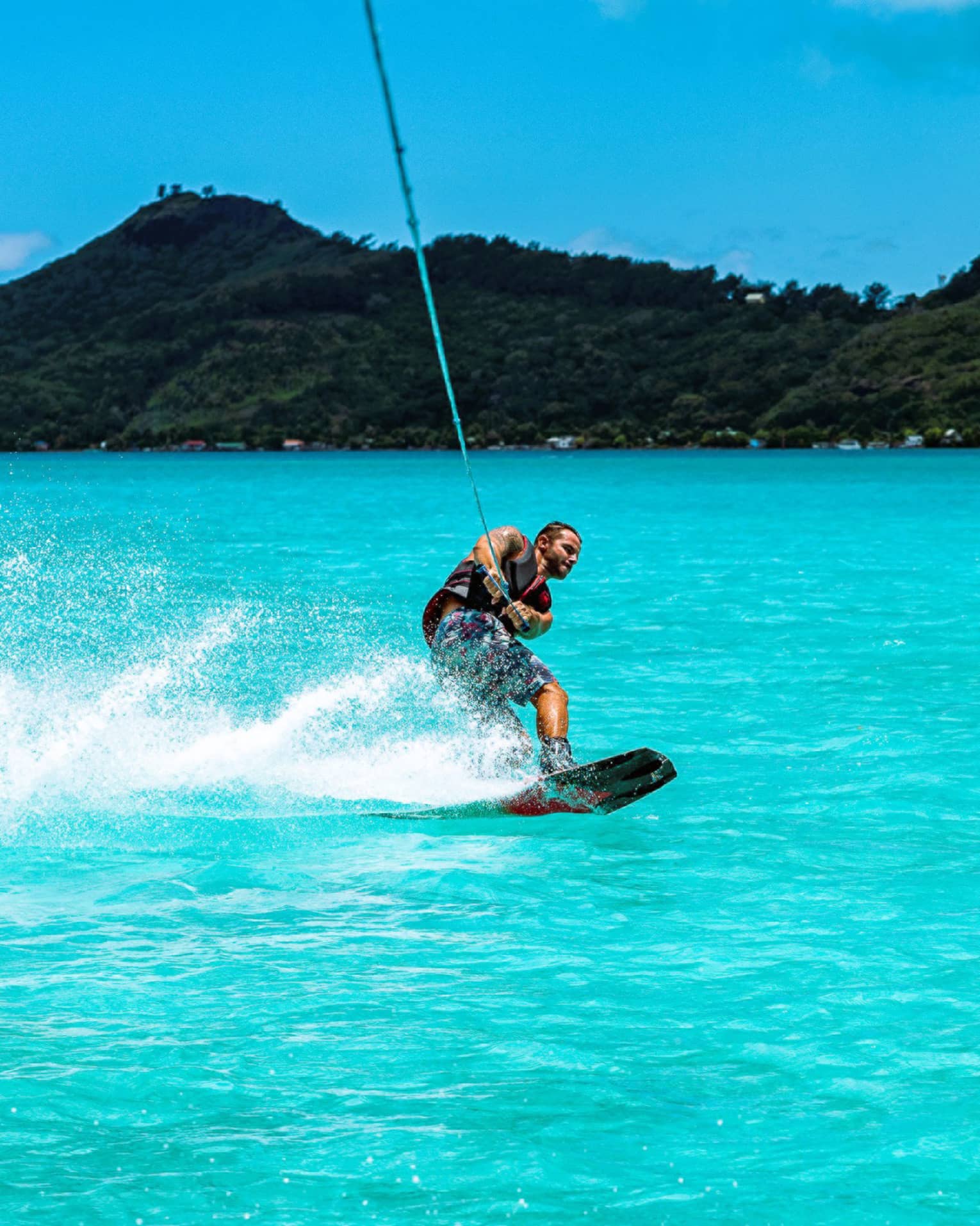 A guest creates a spray of water with their wakeboard as they're towed through the ocean before a backdrop of forested hills.