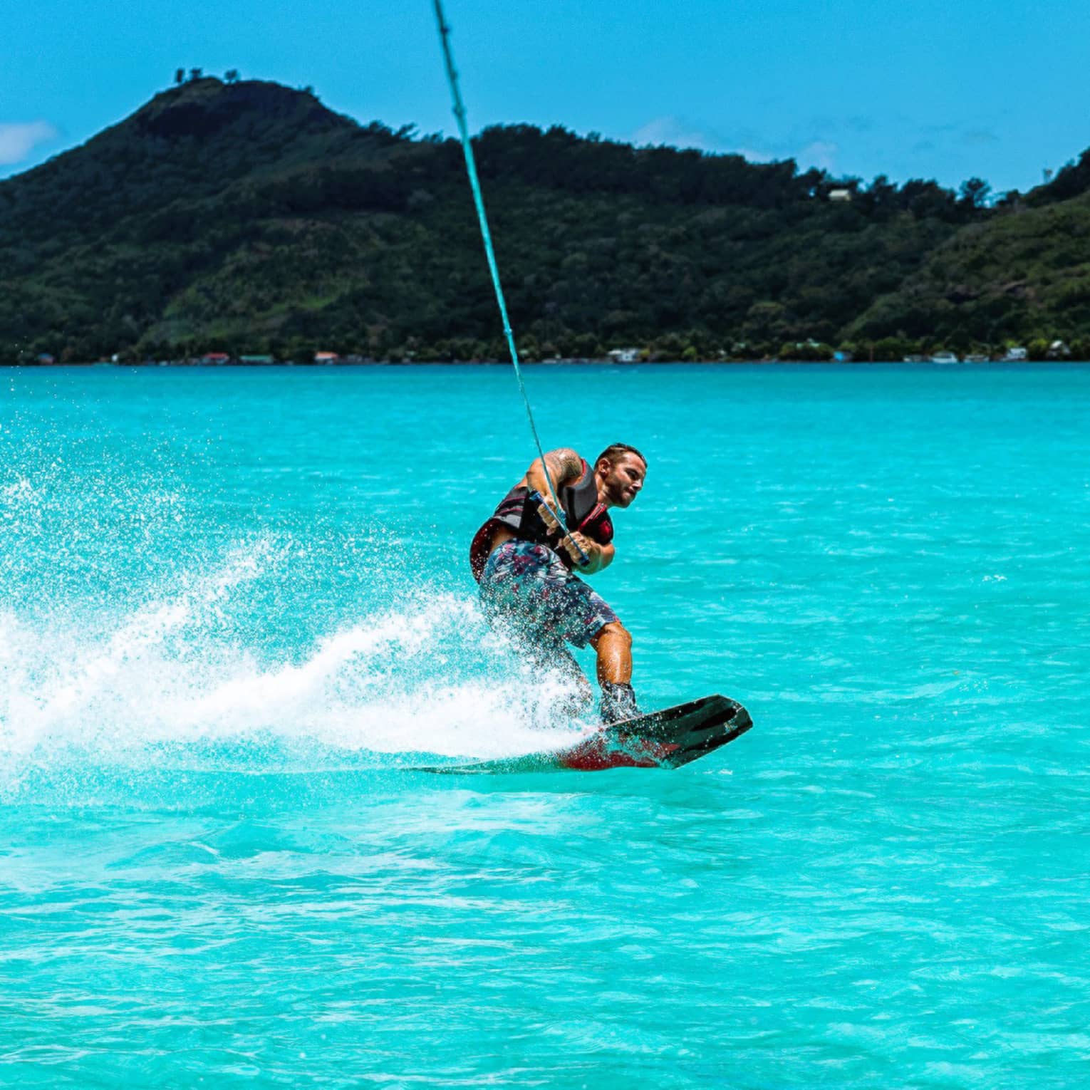A guest creates a spray of water with their wakeboard as they're towed through the ocean before a backdrop of forested hills.